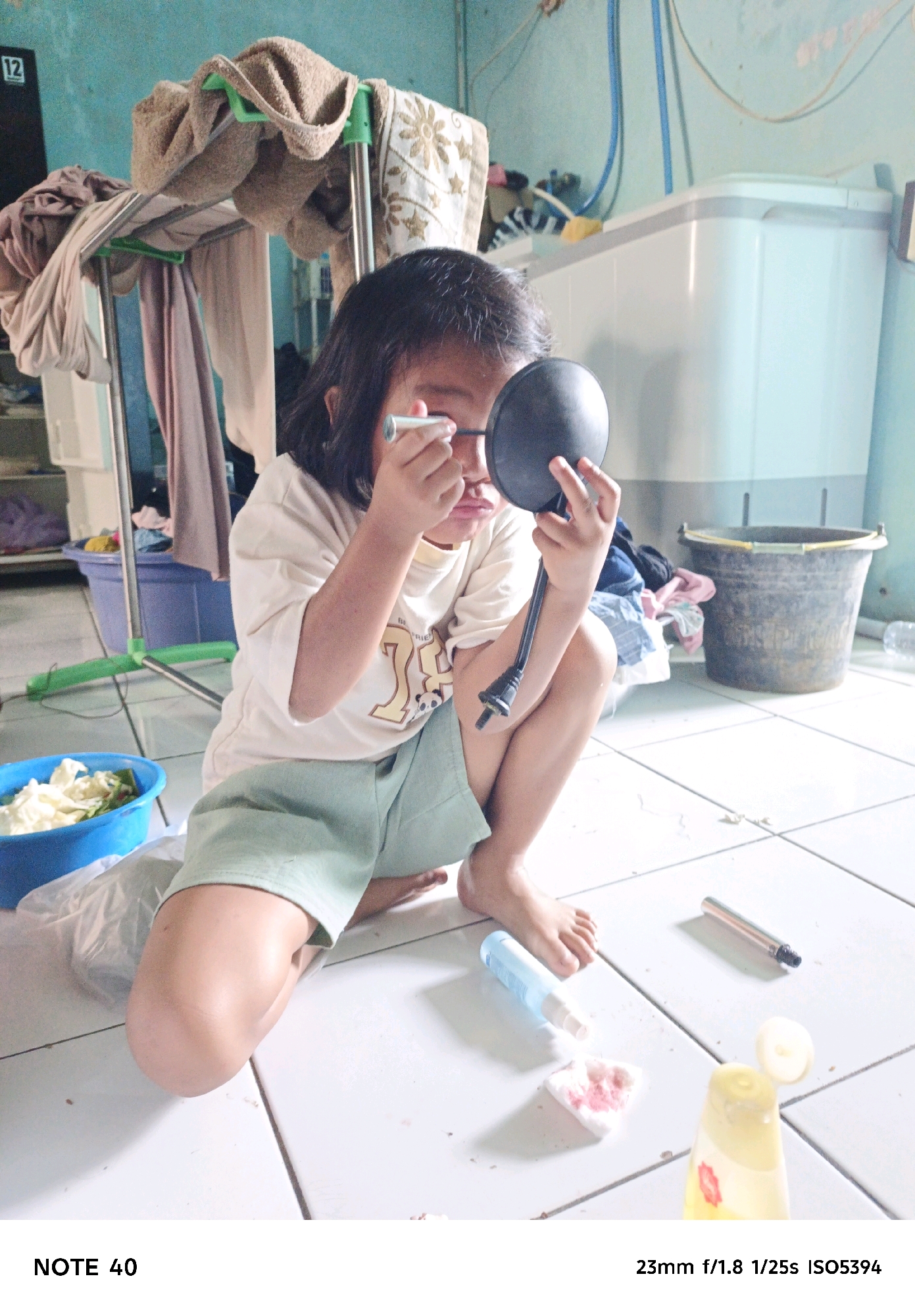 A young girl, possibly Southeast Asian, is squatting on a tiled floor and appears to be applying makeup to her eye with a wand. She is holding a compact mirror in her other hand. She is wearing a light-colored t-shirt and light green shorts. The background shows a laundry area with a washing machine, a large dark bucket, and laundry on a drying rack. Various cleaning or personal care items are scattered on the floor around her.