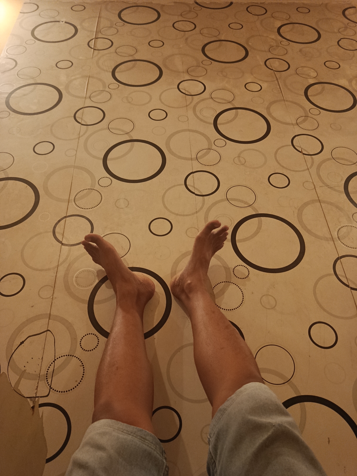 The image shows a low-angle shot of a person's legs and feet on a floor with a circular pattern. The person is wearing light blue denim shorts. The floor is light brown with various sized black and grey circles, some solid and some made of dots. The lighting is warm and appears to be from a single source, casting subtle shadows. The scene is indoors. The pattern on the floor is consistent with flooring found in residential or commercial spaces. There are no other people or objects visible in the frame.