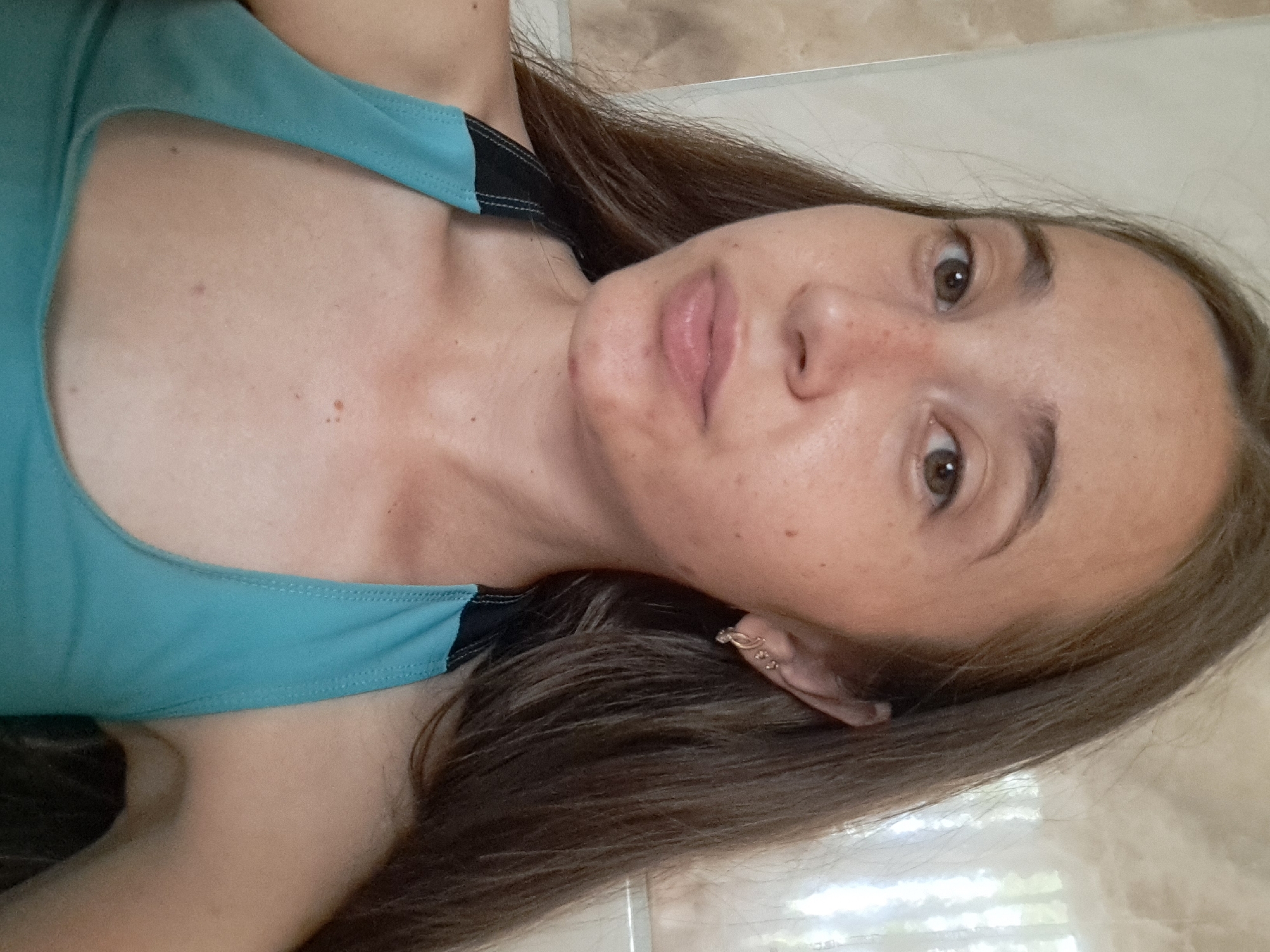A young woman with long, dark brown hair is captured in a close-up selfie, her gaze directed forward. She has light brown eyes and fair skin with freckles scattered across her nose and cheeks. A single, small stud earring is visible in her left ear, and a larger hoop earring with a decorative element is in her right ear. She is wearing a teal-colored athletic top with black straps. The background appears to be a tiled wall, suggesting an indoor setting, possibly a bathroom or shower area. The image is taken from the chest up.