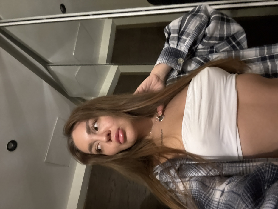 A young woman with long brown hair, wearing a white tube top and a blue and white plaid shirt, poses for a selfie. She is indoors, with mirrored closet doors visible in the background. A silver heart necklace and a Roman numeral tattoo are visible on her chest. Her gaze is slightly off-center, and her lips are plump. A recessed lighting fixture is seen on the ceiling.