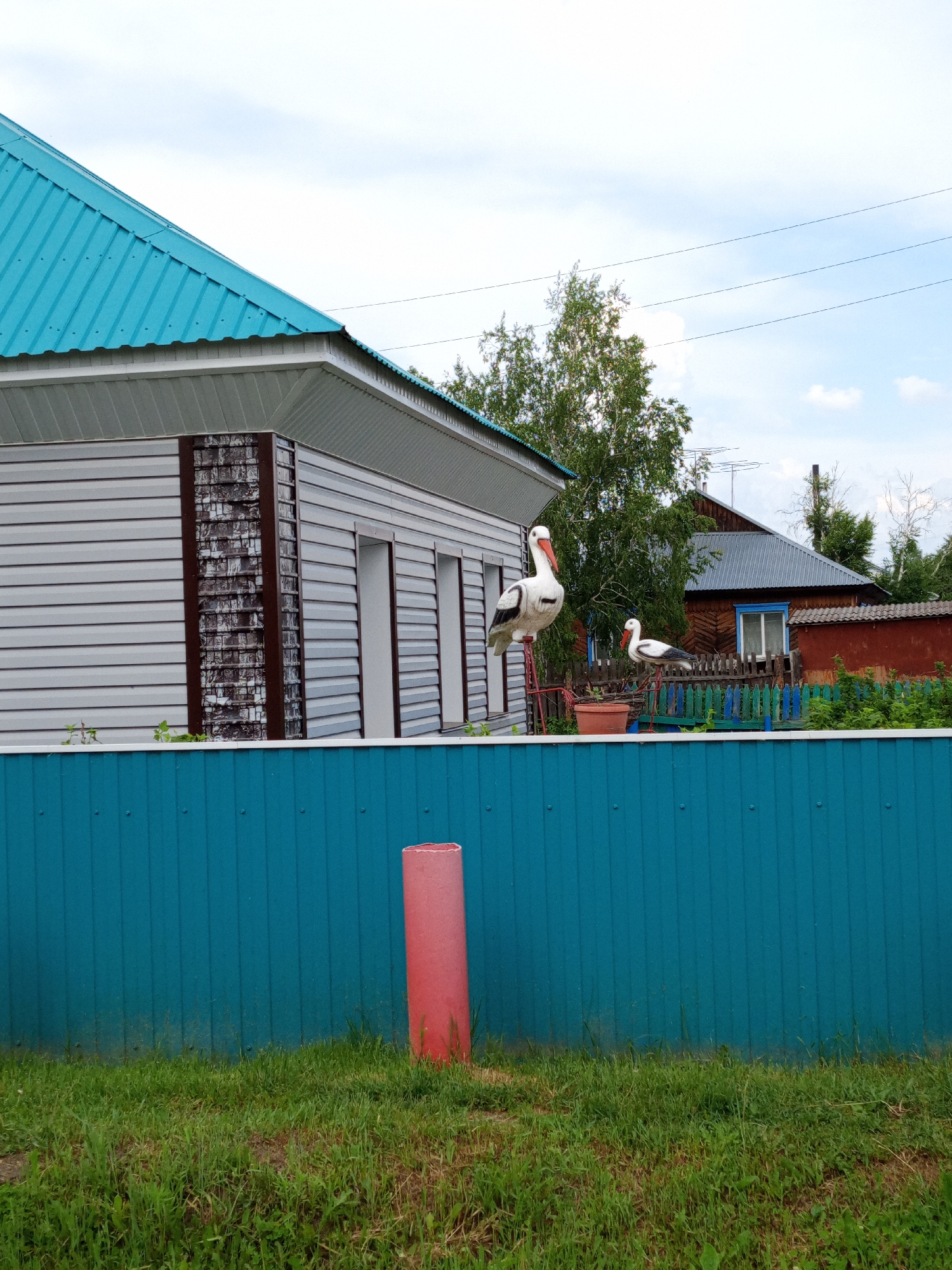 The image depicts a scene in the village of Pankrushikha, Russia. In the foreground, a bright turquoise metal fence runs horizontally across the frame, with a grassy area below and a red cylindrical object positioned to the right of the fence. Behind the fence, a light gray, horizontally-paneled house with a turquoise metal roof is visible. Decorative stone-like elements in brown and dark gray are present on the corner of the house. To the right of this house, two ornamental storks stand on red metal legs, one larger than the other, positioned behind the fence. Further in the background, a traditional wooden Russian house with a gray metal roof and a red section of fence is partially obscured by lush green trees and foliage. The sky is partly cloudy with a light blue hue.