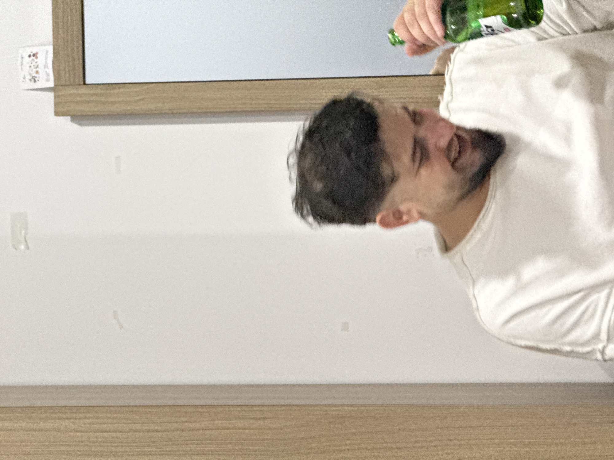The image depicts an indoor scene, likely set in Đà Nẵng, Vietnam, featuring a man in the act of drinking from a green bottle. The man, with dark hair and a beard, is wearing a plain white t-shirt. He is captured mid-action, with his head tilted back and his mouth wide open, suggesting a moment of strong emotion such as laughter, surprise, or enthusiastic consumption.
His right hand holds the green bottle, which has a white label featuring an unclear Asian script, likely Korean characters, typical of a soju bottle. A mirror with a light wooden frame is visible behind and above the man, partially reflecting the plain white wall. In the top-left corner, a small, blurry white tag with a faint colorful pattern is tucked into the mirror's frame. The background wall is plain white, showing a few indistinct smudges. Below the wall and the man, a wooden surface, possibly a shelf or part of a piece of furniture, spans the width of the frame. The lighting is soft, and the image has a slightly grainy quality. The time of day is not discernible, but the candid nature of the shot suggests an informal moment of enjoyment.