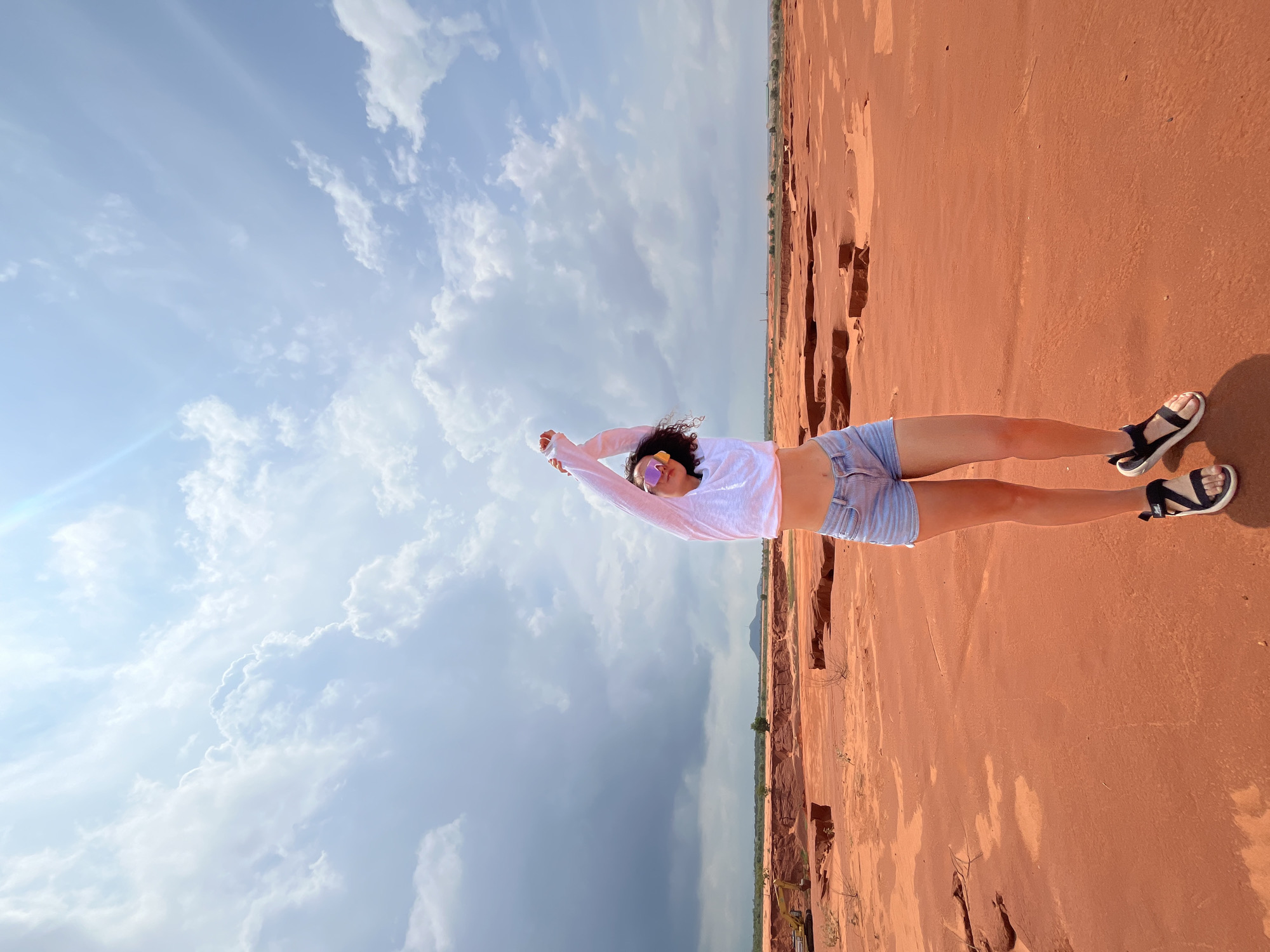 A young woman with curly dark hair and sunglasses stands in a desert-like landscape, stretching her arms up towards a bright sky filled with scattered clouds. She is wearing a white crop top and light blue denim shorts. The ground is a reddish-orange hue, with visible erosion patterns and some dry brush. In the distance, a yellow excavator is partially visible, suggesting ongoing activity or past development in the area. The background shows a somewhat barren landscape with hills and scattered trees under a wide expanse of sky. This scene is characteristic of the red sand dunes and sand quarries found in Mui Ne, Vietnam, a popular tourist destination known for its unique geological formations.