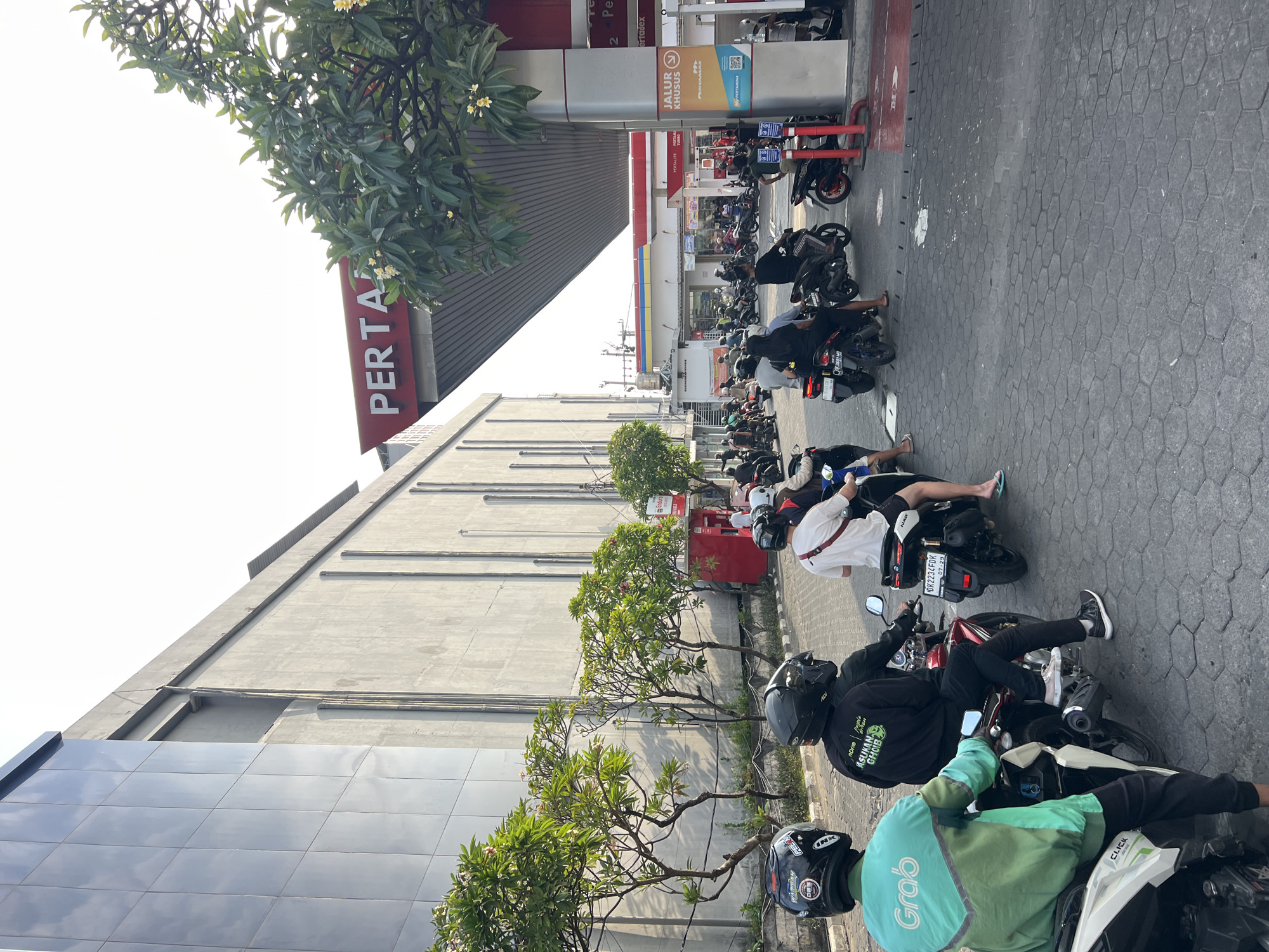 This is a medium shot, eye-level, outdoor photograph taken in Kuta, Indonesia. The scene depicts a busy Pertamina gas station with numerous motorcycles and their riders. The architecture is modern, with a large, grey, concrete building on the left featuring a section with reflective, square panels. A prominent red and white Pertamina canopy is overhead, advertising "PERTAMAX." Several motorcycles are lined up, suggesting a queue for fuel. A Grab driver in a green and light blue uniform is visible in the foreground on the left, mounted on a motorcycle. Other individuals on motorcycles are also present, some wearing helmets. In the background, there are trees with green foliage and some flowering plants. The ground is paved with dark hexagonal tiles. The sky is bright and overcast. The overall impression is one of daily activity at a fuel station in a commercial area.