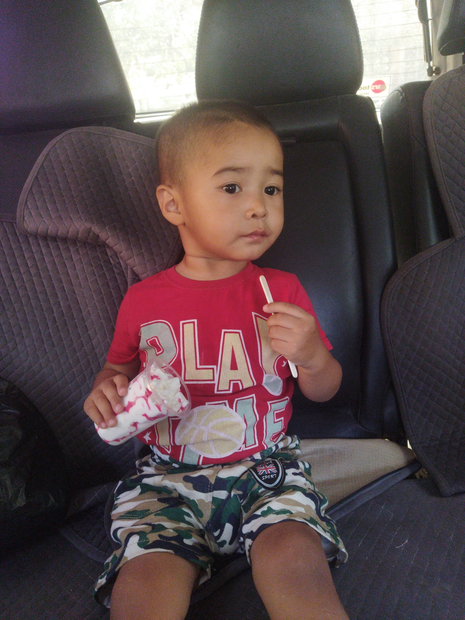 A young boy sits in the back seat of a car, holding a plastic cup with a dessert and a spoon. He is wearing a red t-shirt with a graphic print and camouflage shorts. The car seats are black and upholstered with a quilted pattern. The background shows car windows with a blurred view of trees and a sticker on the window. The location is specified as Mailuu-Suu City, Kyrgyzstan.