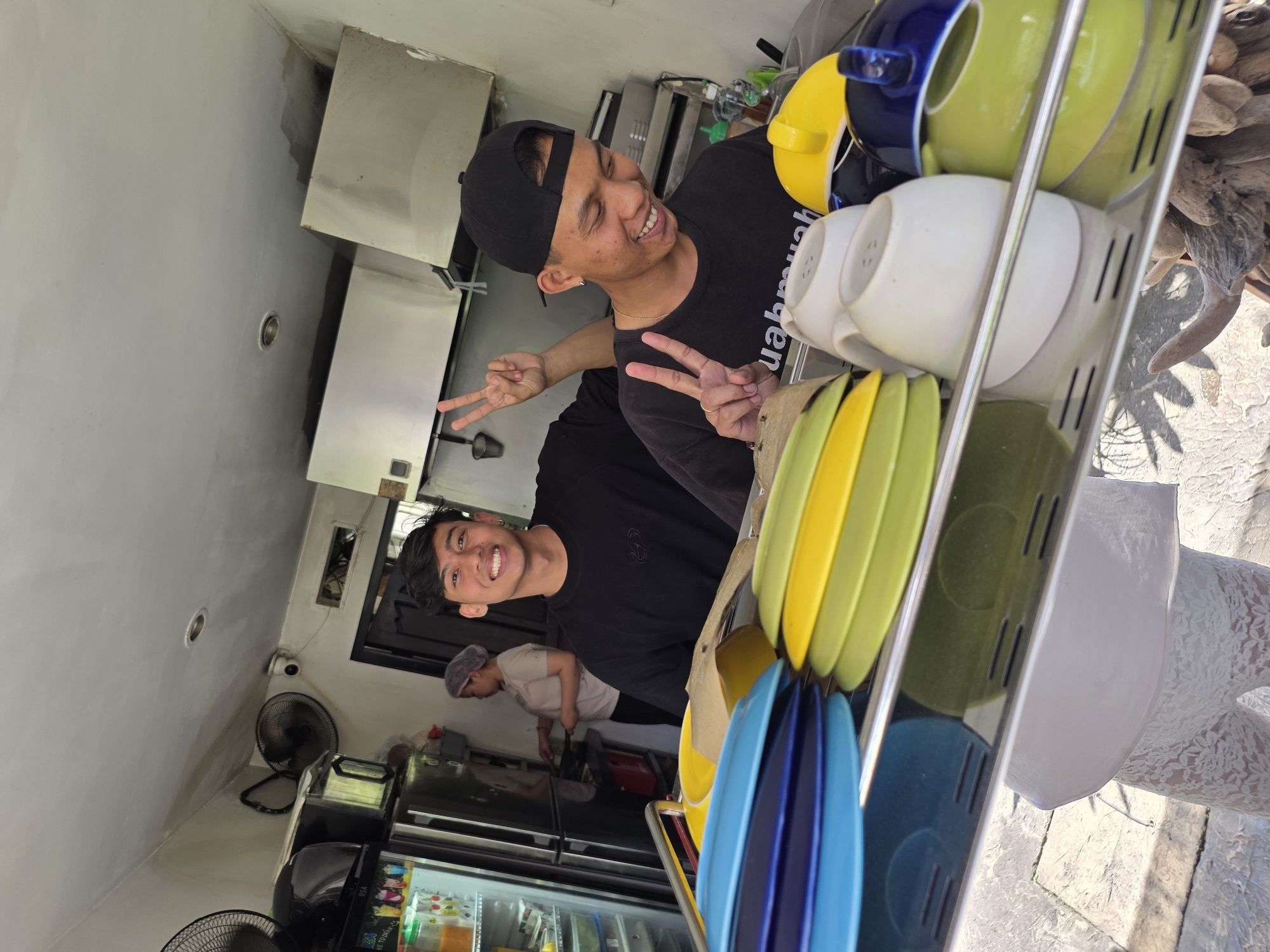 Two young men stand near a stainless-steel rack holding brightly colored mugs and plates. The first man, wearing a black baseball cap and t-shirt, flashes a peace sign and smiles. The second man, behind the first, also smiles and flashes a peace sign. Behind them, an individual wearing a head covering is visible in the background, near a refrigerator filled with beverages. A CCTV camera is mounted on the wall. An electric fan sits on the floor. A stainless steel cabinet is visible in the upper-left corner. The exterior background shows a white surface with a pattern and what appears to be vegetation or driftwood. The location is North Kuta, Indonesia.