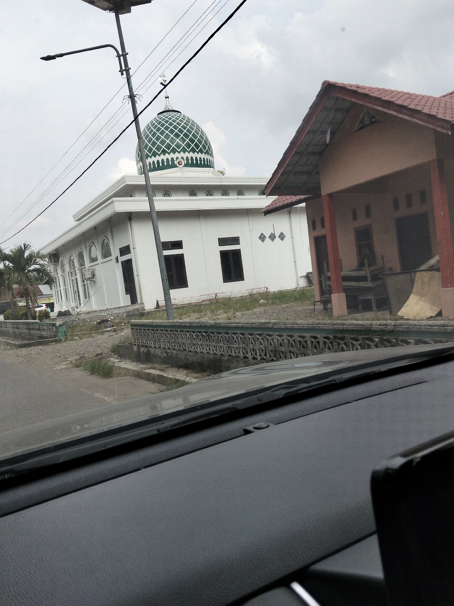 The image depicts a street view in Awegeutahpaya, Indonesia, focusing on a white mosque with a green dome and an adjacent beige building. The scene appears to be captured from inside a vehicle, as the lower part of the image shows a car's dashboard and windshield. A street lamp and power lines are prominent in the foreground, running diagonally across the frame. The mosque, characterized by its arched windows and a detailed dome pattern, stands to the left, while the other building, with a red tiled roof and open-fronted sections, is on the right. A decorative fence runs between the road and the buildings. The environment is overcast, with a scattering of vegetation and debris visible around the structures.