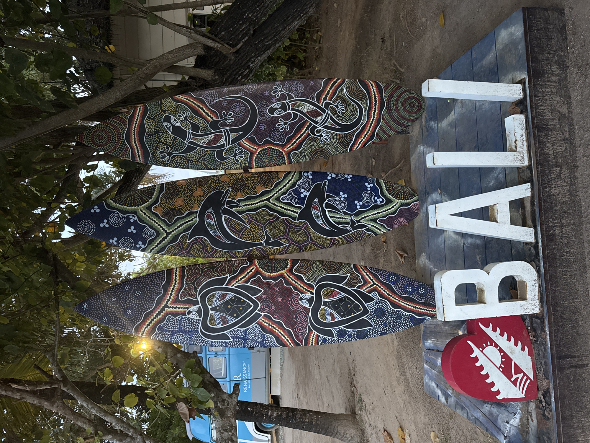 Three surfboards are stacked vertically against a tree trunk, angled to the left. Each surfboard is painted in an Aboriginal-style dot art design featuring animals: a gecko on the top board, dolphins on the middle board, and turtles on the bottom board. To the right of the surfboards, the word "BALI" is spelled out vertically in large, white block letters attached to a weathered wooden board. A red heart shape with a white sun and wing design is affixed below the "B". The scene appears to be outdoors, with a dirt or sandy ground surface visible, and branches of a tree framing the top and left side of the composition. A blue van with the text "Renaissance Bali Uluwatu Resort" on the side is visible in the background. This scene is located in Kuta Selatan, Indonesia.