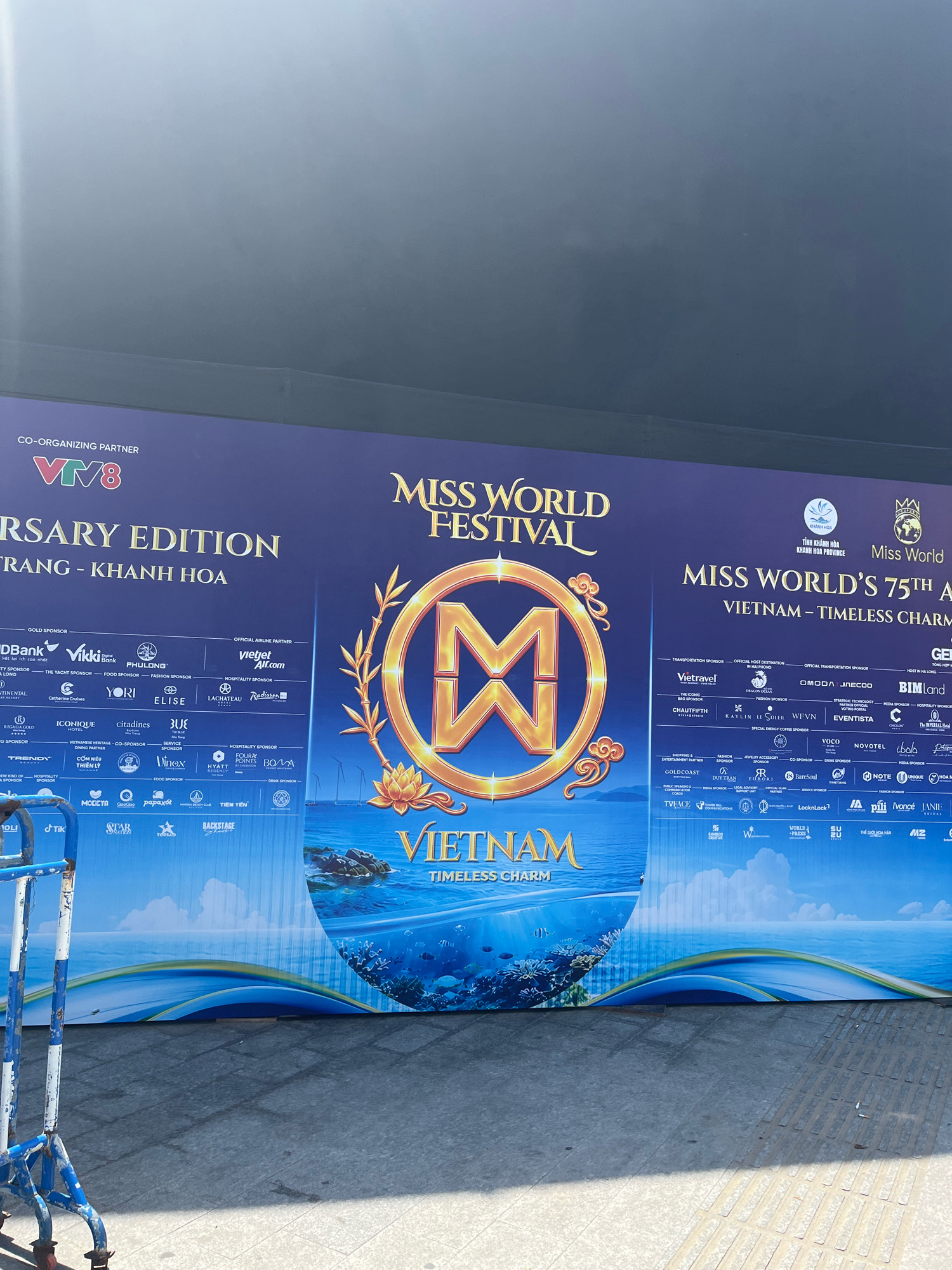 The image shows a large banner for "MISS WORLD FESTIVAL" and "MISS WORLD'S 75TH ANNIVERSARY EDITION VIETNAM – TIMELESS CHARM". The banner features a prominent golden "M" logo, an underwater scene with coral and fish, and numerous sponsor logos. The banner is positioned outdoors, with a cobblestone-like pavement in the foreground and a railing of a blue and white cart on the left side. In the background, the outline of wind turbines can be seen against a bright sky, suggesting an open, possibly coastal area. The overall impression is of an event advertisement displayed in a public space in Nha Trang, Vietnam.