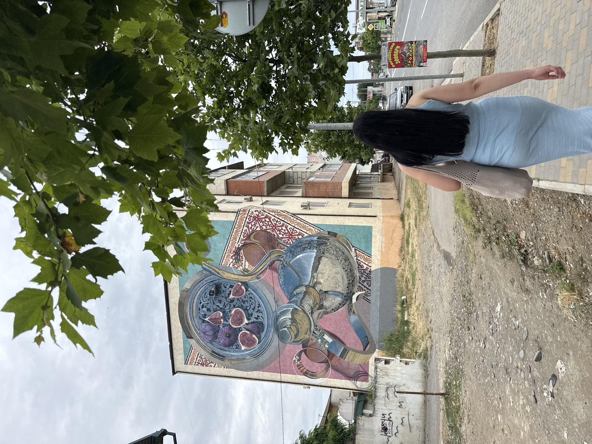 A woman with long, dark hair, wearing a light blue knitted dress and carrying a light grey tote bag, walks away from the camera down a sidewalk. To her left, a tall building features a large, colorful mural on its side depicting a silver teapot, a tray of figs, and intricate patterns. The mural is framed by a blue border. The building itself is multi-story with balconies. To the right of the woman is a street with cars and a sidewalk. A red and yellow sign is attached to a pole near the sidewalk. The environment is urban, with some greenery from trees and scattered debris on the ground. The sky is overcast. The building with the mural is located in Derbent, Russia.