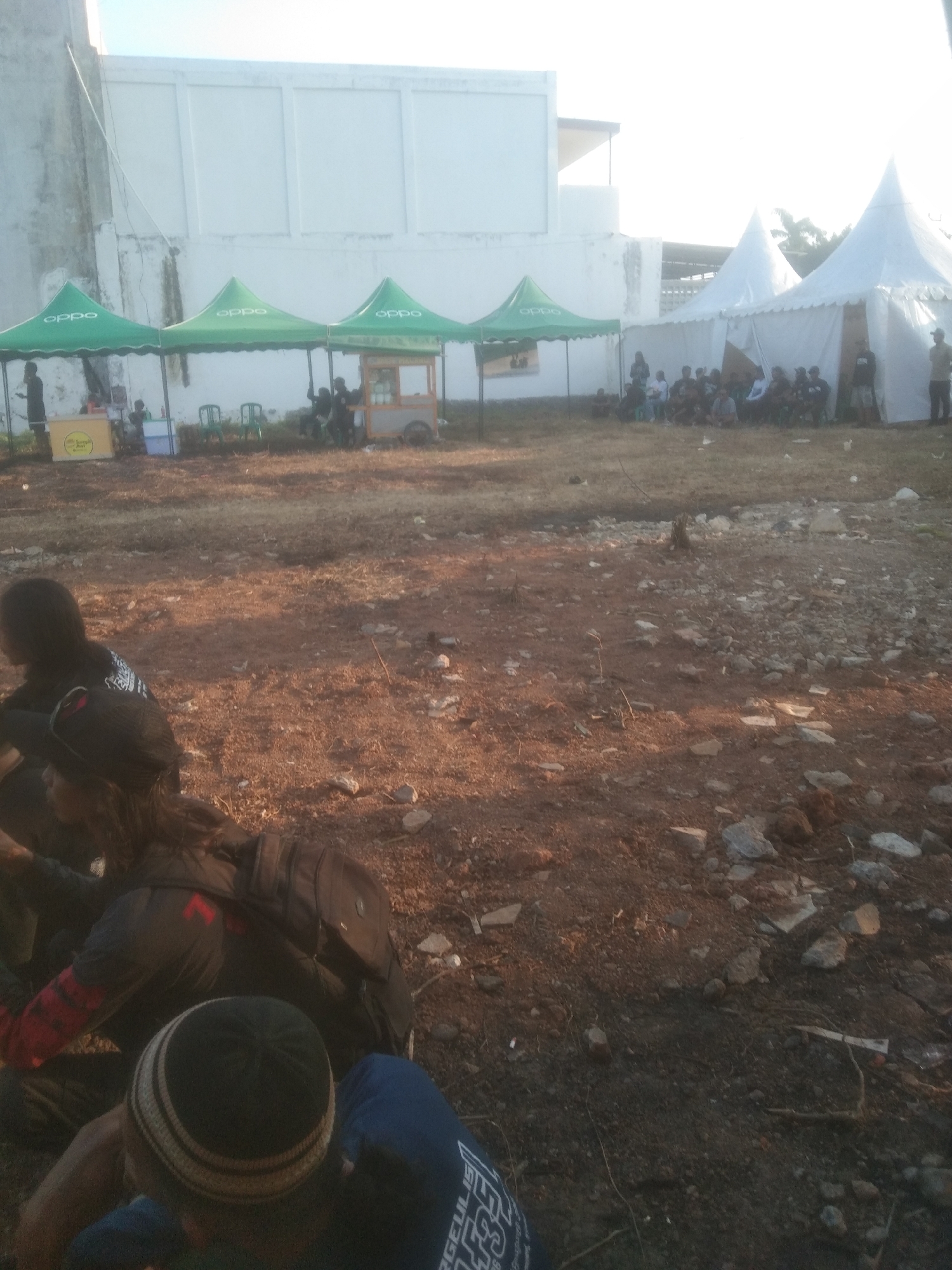 In Kecamatan Haurgeulis, Indonesia, an outdoor scene depicts a gathering of people under several large white tents and green tents. In the background, a white building with a flat roof is visible. In the foreground, the ground is covered in dry dirt and scattered rocks. Several individuals are seated or crouched on the ground. Some are wearing casual clothing, including a person with a black beanie and another with a black backpack. There are also some vendor stalls set up under the green tents, one with a cart that appears to be selling food or drinks. A group of people are gathered around the white tents, some standing and some sitting. The overall impression is of a casual outdoor event or market.