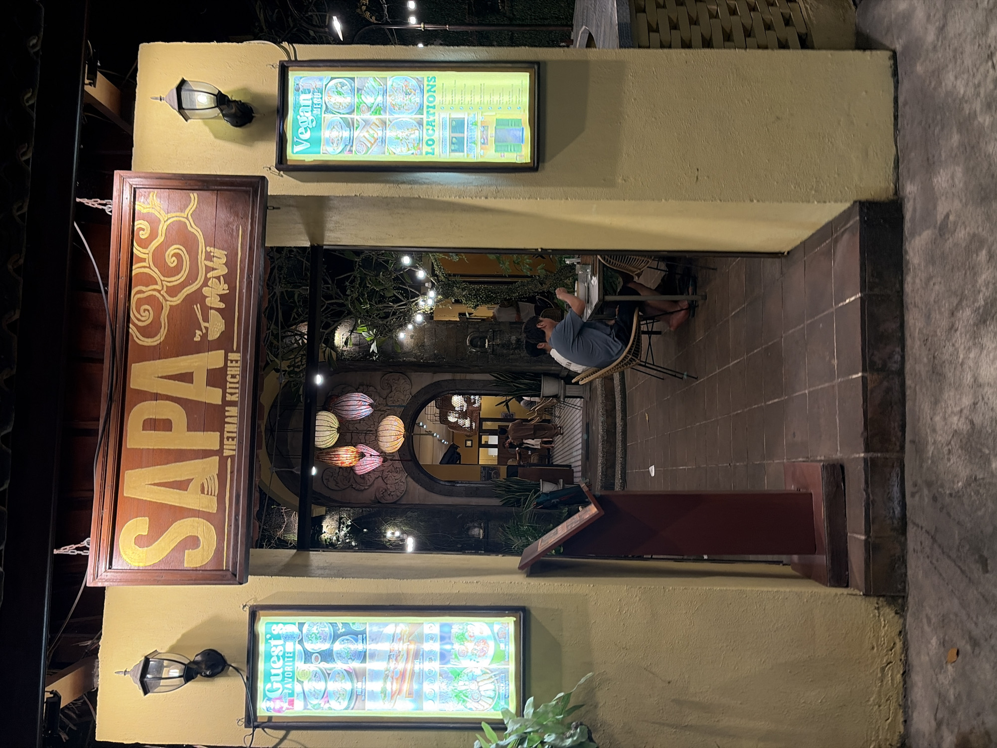 The image depicts the entrance to a restaurant called "SAPA VIETNAM KITCHEN" in Ubud District, Indonesia. The entrance is flanked by a textured yellow wall and illuminated by traditional black lanterns. Two illuminated signs are mounted on the wall: one on the left is labeled "Guest's Favorite" and displays images of Vietnamese dishes, while the one on the right is labeled "Vegan MENU" and also shows food items. A wooden menu stand is positioned in front of the entrance. Inside the restaurant, visible through an archway, is a dimly lit courtyard decorated with colorful lanterns and string lights. Two individuals are seated at a table in the courtyard, appearing to be dining. The overall ambiance suggests a tropical evening setting.