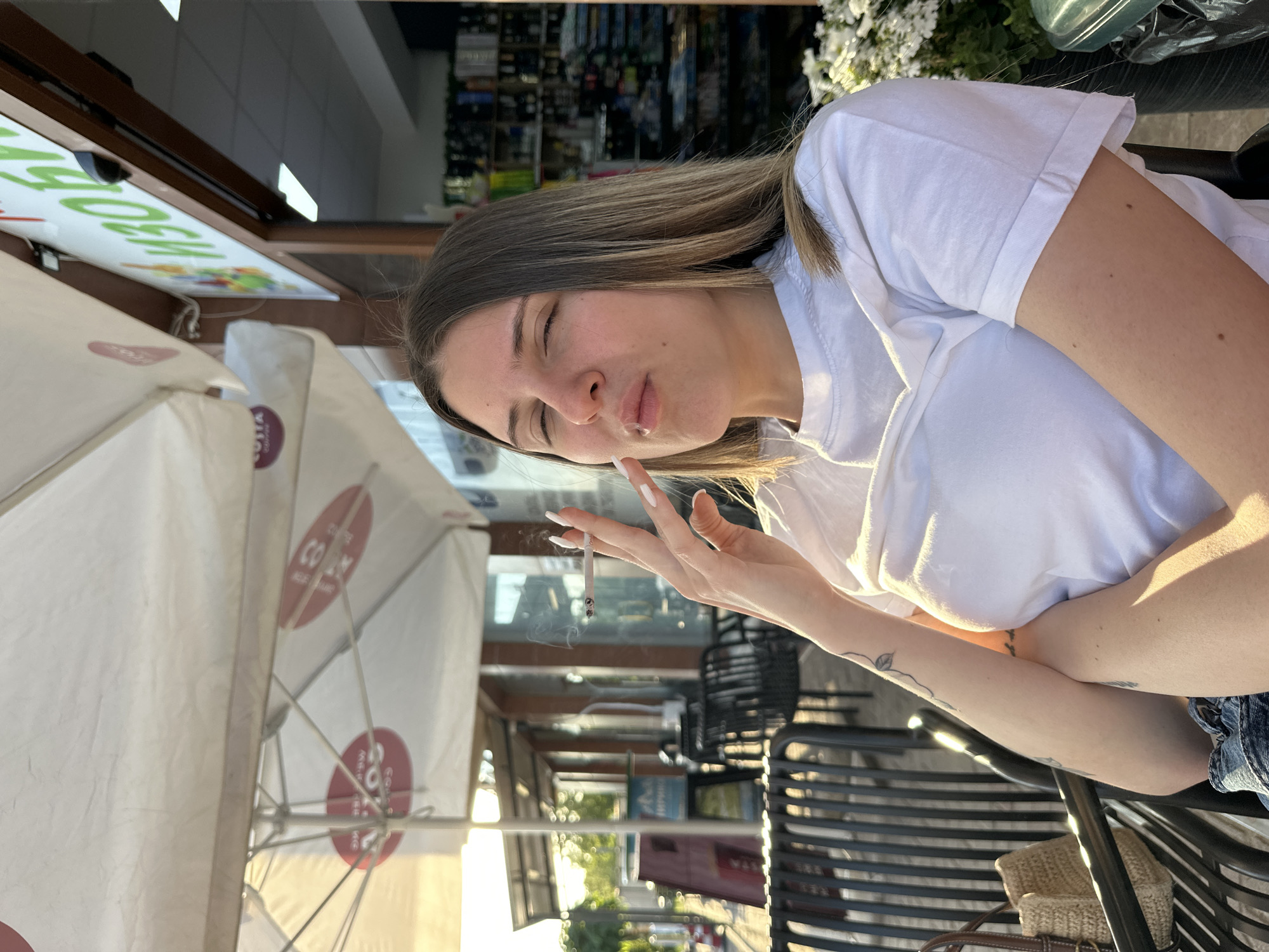 A young woman with light brown hair is sitting outdoors at a cafe. She is wearing a white t-shirt and holding a lit cigarette. The cafe has white umbrellas with red writing on them, a storefront with a sign in a foreign language, and metal chairs. The background shows a street scene. The location is likely Plovdiv, Bulgaria.