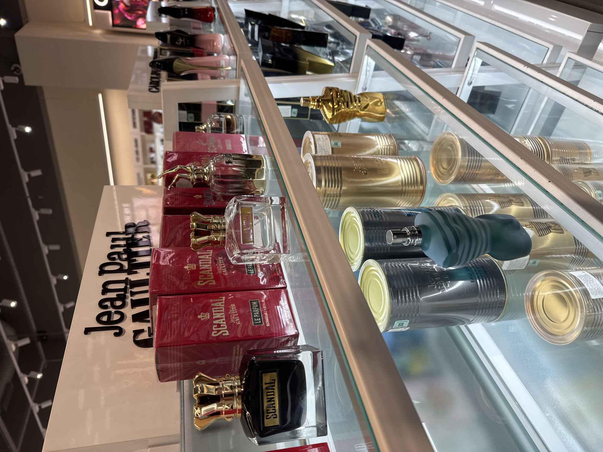 This is a close-up, upward-angled photograph of a retail display featuring a variety of perfumes from the Jean Paul Gaultier and Carolina Herrera brands. The products are arranged on glass shelves within a brightly lit display case. The bottles of perfume are predominantly in red and gold packaging, alongside the 'can' style packaging associated with Jean Paul Gaultier. The camera is positioned to highlight both the products on the shelves and the reflections within the glass of the display case. Based on the brand names on display, it is likely this photograph was taken in a department store or duty-free shop in Yerevan, Armenia. The text on the packaging is mainly in English and possibly French.