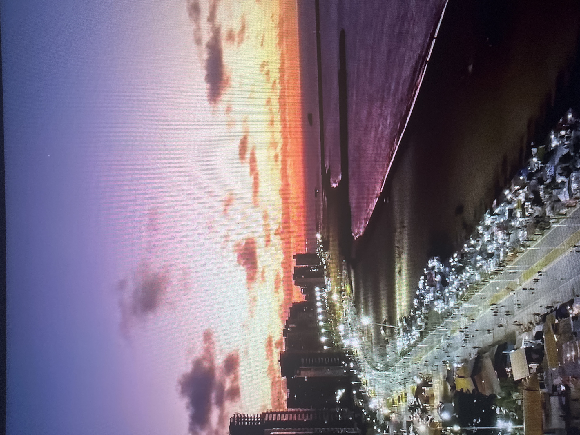 An aerial, long-shot perspective captures a city coastline at twilight. The sky transitions from a deep violet at the top to a soft pink and orange gradient near the horizon, where the sun has set. Scattered clouds, tinged with pink and orange, are visible in the sky.
Below the sky, a cityscape lines the coast. Numerous tall, modern buildings are silhouetted against the colorful sky. A well-lit promenade follows the curve of the beach, with rows of streetlights illuminating the pathway. Many people are visible walking along the promenade, and what appear to be stalls or temporary structures are set up along its edge, also illuminated.
The beach itself is dark, appearing to be made of sand. The ocean is a deep, shimmering purple, reflecting the twilight sky. A long, dark breakwater extends into the water from the right side of the frame, with another, shorter breakwater or pier visible closer to the shore. A small, dark object, possibly a boat or a buoy, is visible in the distance on the water. The overall scene depicts a lively urban coastline transitioning from day to night.