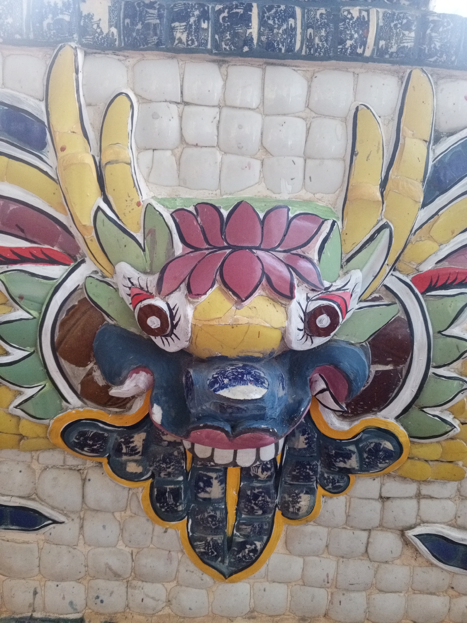 This is a close-up, eye-level, vertical, full shot of a decorative dragon head mosaic on the exterior wall of the Long Hai Temple in Xuan Thanh, Vietnam. The mosaic is made of colorful ceramic tiles and depicts a mythical dragon with yellow horns, green and red foliage, a blue face, and a red and pink mouth with visible teeth. The dragon's eyes are circular with brown irises and white pupils. The background consists of white square tiles. The mosaic is weathered and shows some signs of age and damage.