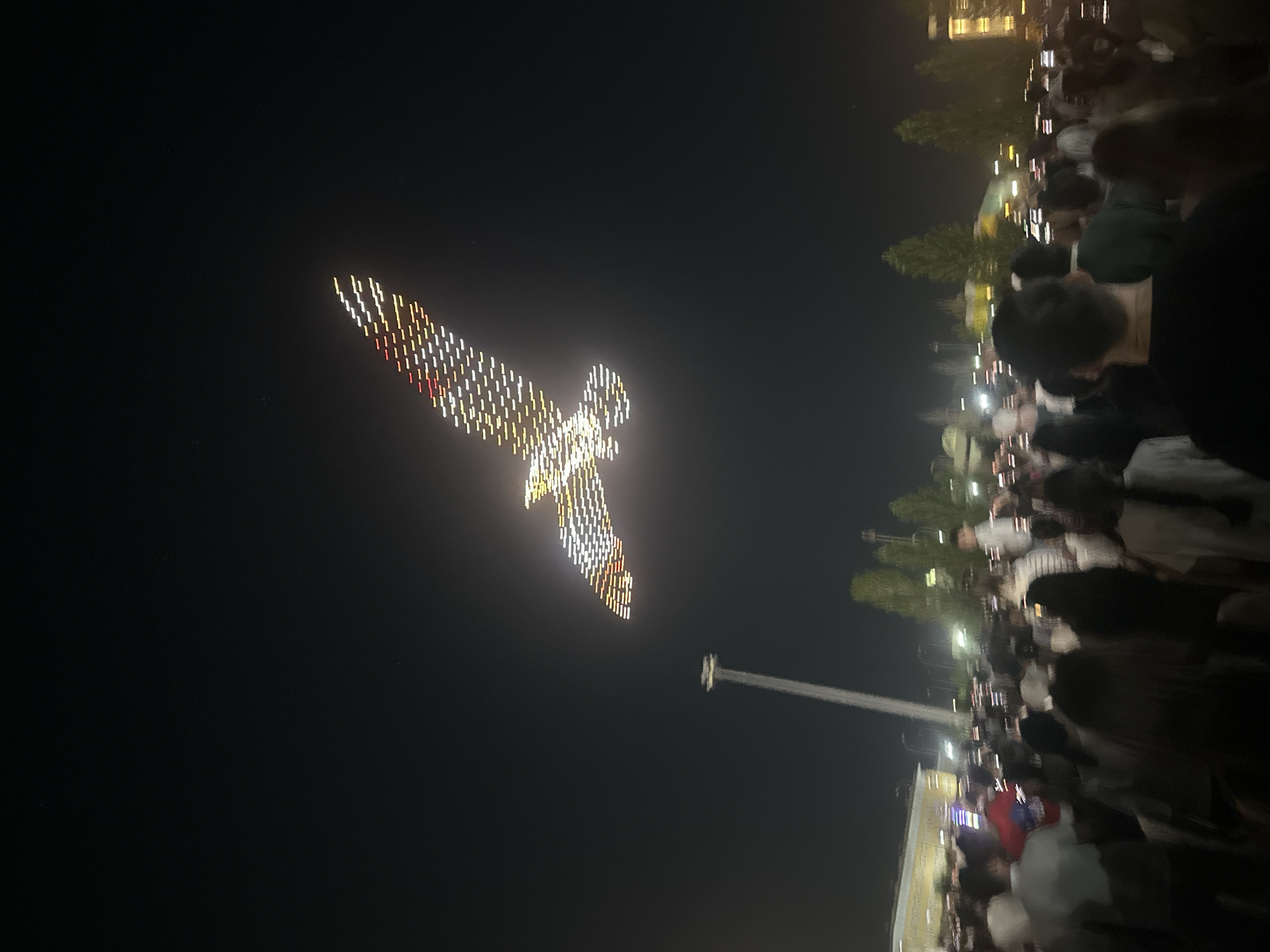 The image captures a drone light show in the city of Oral, Kazakhstan, at night. A large formation of drones is in the sky, creating the shape of a bird with outstretched wings, illuminated by white and red lights. Below the drone display, a large crowd of people is gathered, many holding up their phones to capture the spectacle. The background shows the urban environment of Oral with some buildings and trees visible, as well as a tall structure resembling a tower. The overall scene suggests a public event or celebration taking place in the city.