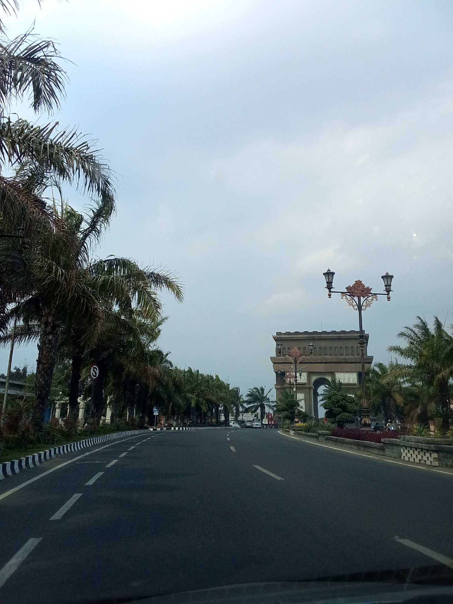 The image shows a street scene in Ngasem, Indonesia, with a grand archway structure in the background. On the left side of the road, a row of tall palm trees lines the street, with their fronds appearing somewhat dried and sparse. The road itself is paved and has white dashed lane markings. A white and black striped curb separates the road from the sidewalk. In the distance, a large, ornate archway, possibly a monument or gateway, stands prominently. It has a squared-off top and a large arched opening. Decorative lampposts with multiple lights are visible in front of and near the archway. More palm trees and greenery are scattered around the monument and along the right side of the road. The sky is overcast with a pale, diffused light. A few vehicles, including cars and a motorcycle, are present on the road, suggesting moderate activity. The overall impression is of an urban thoroughfare with significant architectural elements and tropical vegetation.