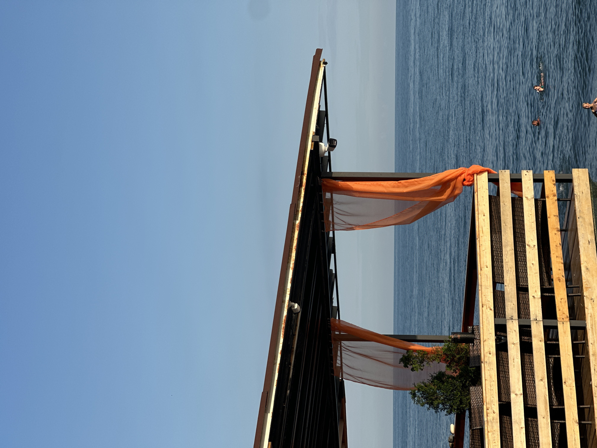 The image depicts an outdoor seating area with a wooden railing and brown wicker chairs, overlooking the sea. Orange translucent curtains are hung from the overhead structure, providing shade. In the background, several people are swimming or floating in the blue water. The sky is clear and blue. The location appears to be a beachside cafe or restaurant in Khostinskiy, Russia.