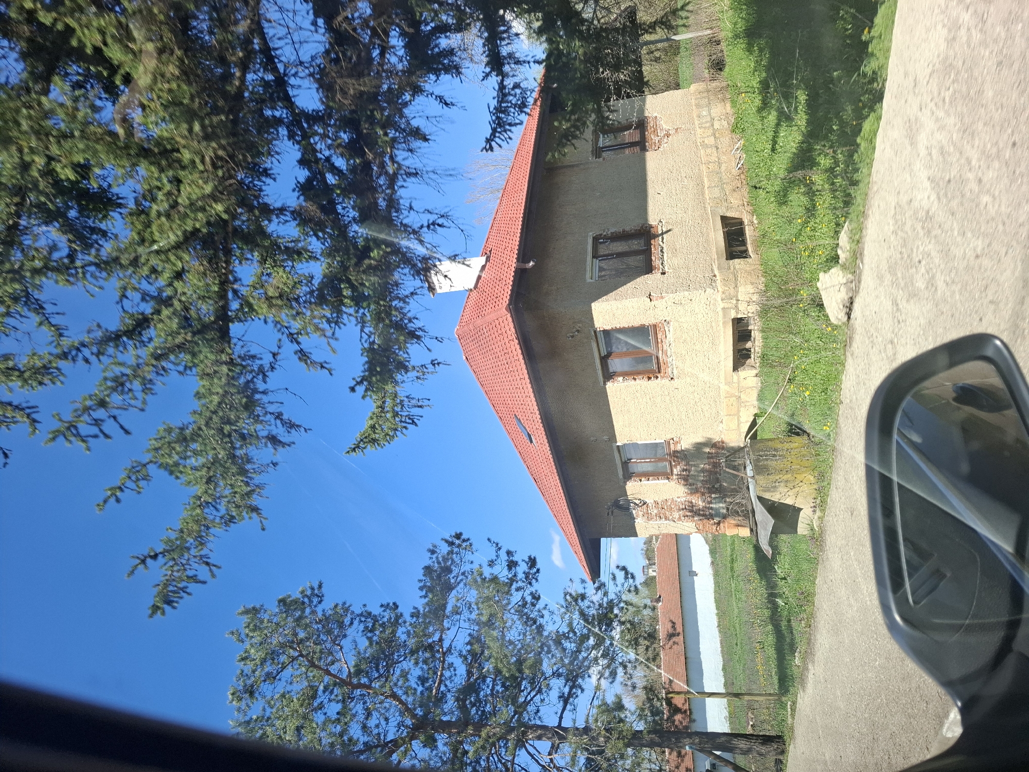 The image shows a building with a red tiled roof, possibly residential, viewed from a car window. Part of a side mirror is visible. The building appears to be in a state of disrepair, with some brickwork exposed. There are trees in the foreground, obscuring the sky. The location is Studeno Buche, Bulgaria, based on prior context. The road and vegetation adjacent to the building are visible. No people are identifiable in the image.