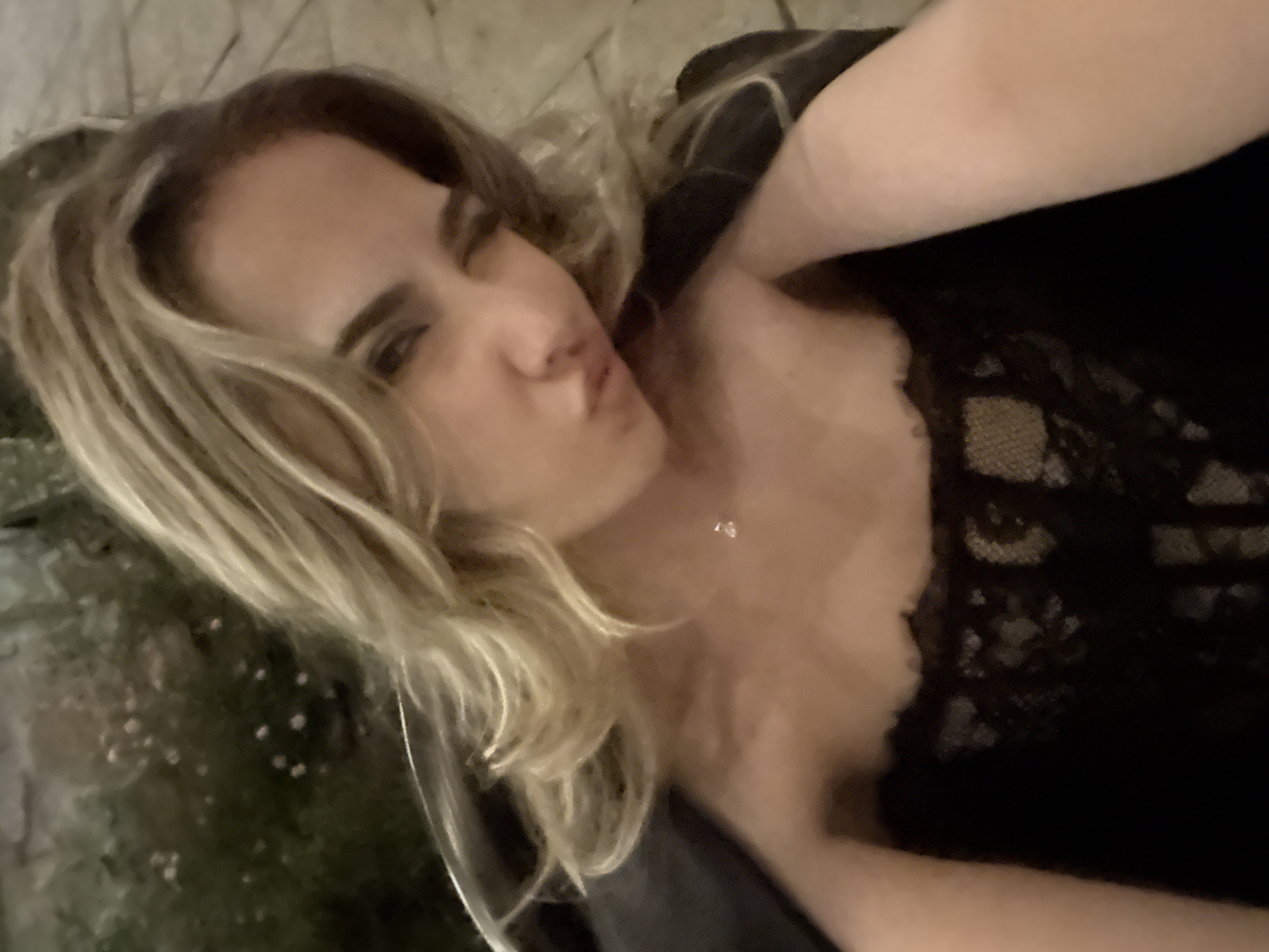 A woman with blonde hair is lying down, wearing a black lacy top. She has light skin, dark eyebrows, and is making a kissing face. A thin necklace is visible on her neck. Behind her, there appears to be greenery and possibly an outdoor stone wall or surface. The specific location of Kocherinovo, Bulgaria, is not discernible from the image.