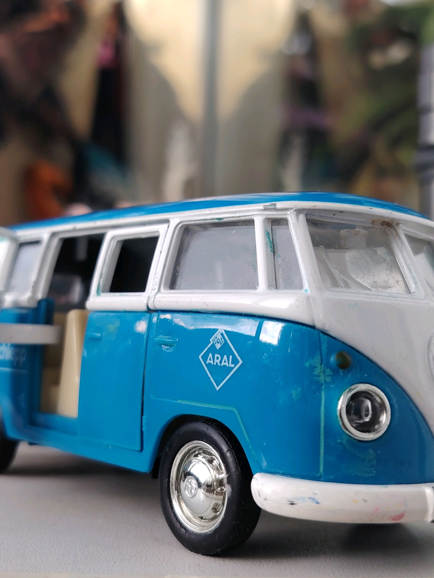 A close-up, low-angle, outdoor shot features a blue and white toy van, specifically a Volkswagen Type 2, positioned on a neutral-colored surface. The toy van is depicted in a side profile, with its front end angled slightly towards the viewer. The van's body is primarily blue, with a white roof and white trim around the windows and front bumper. A diamond-shaped logo with the text "ARAL" is visible on the side of the blue section, near the front door. The left front door of the van is ajar, revealing a cream-colored interior. The toy shows signs of wear, with scuffs and paint marks visible on its surface, particularly on the blue paint. The front wheel is chrome and black, with a silver hubcap. The background is blurred and indistinct, suggesting an out-of-focus environment. There are no people present in the image, and no discernible elements definitively place the scene in Chaikovsky, Russia.