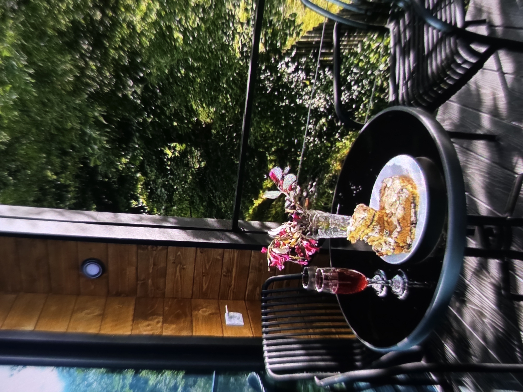 The image depicts an outdoor seating area with a table and two chairs, set against a backdrop of lush green trees. On the black, circular table, there are two glasses filled with red liquid, a vase containing pink and white flowers, and a plate with a slice of cake. The table is situated on a wooden deck. Behind the table and chairs, a wooden-paneled wall features a light fixture and an electrical outlet. The scene is bathed in sunlight, casting shadows on the deck. The general impression is that of a peaceful outdoor setting, likely a balcony or patio, with a refreshing drink and a slice of cake. The specific location in Dubno, Ukraine, is not discernible from the image itself.