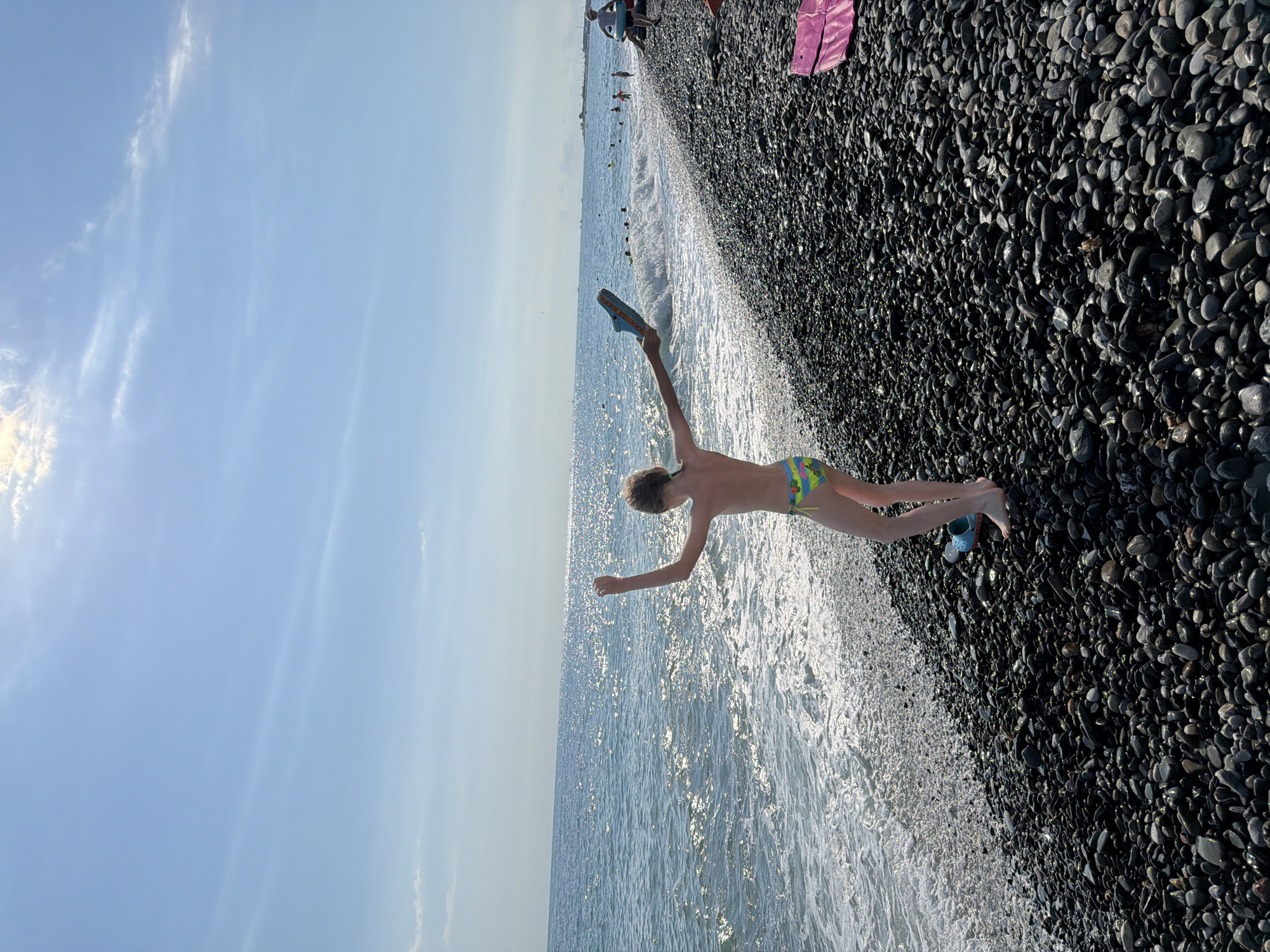 A young person in swim trunks is captured in mid-air, jumping towards the sea on a pebbled beach. The person is on the right side of the frame, their arms extended and legs bent, with one shoe dangling from their raised hand. The sea is to the left, with gentle waves washing onto the shore, creating a shimmering effect from the sunlight. The beach is covered in dark, smooth pebbles. In the background, other beachgoers can be seen near the water's edge, and some are swimming. The sky is a clear, light blue with a few wispy clouds. The sun is shining brightly, suggesting it is daytime and likely a warm day, ideal for a beach outing. The scene evokes a feeling of carefree joy and summer fun. No visible text is present in the image.