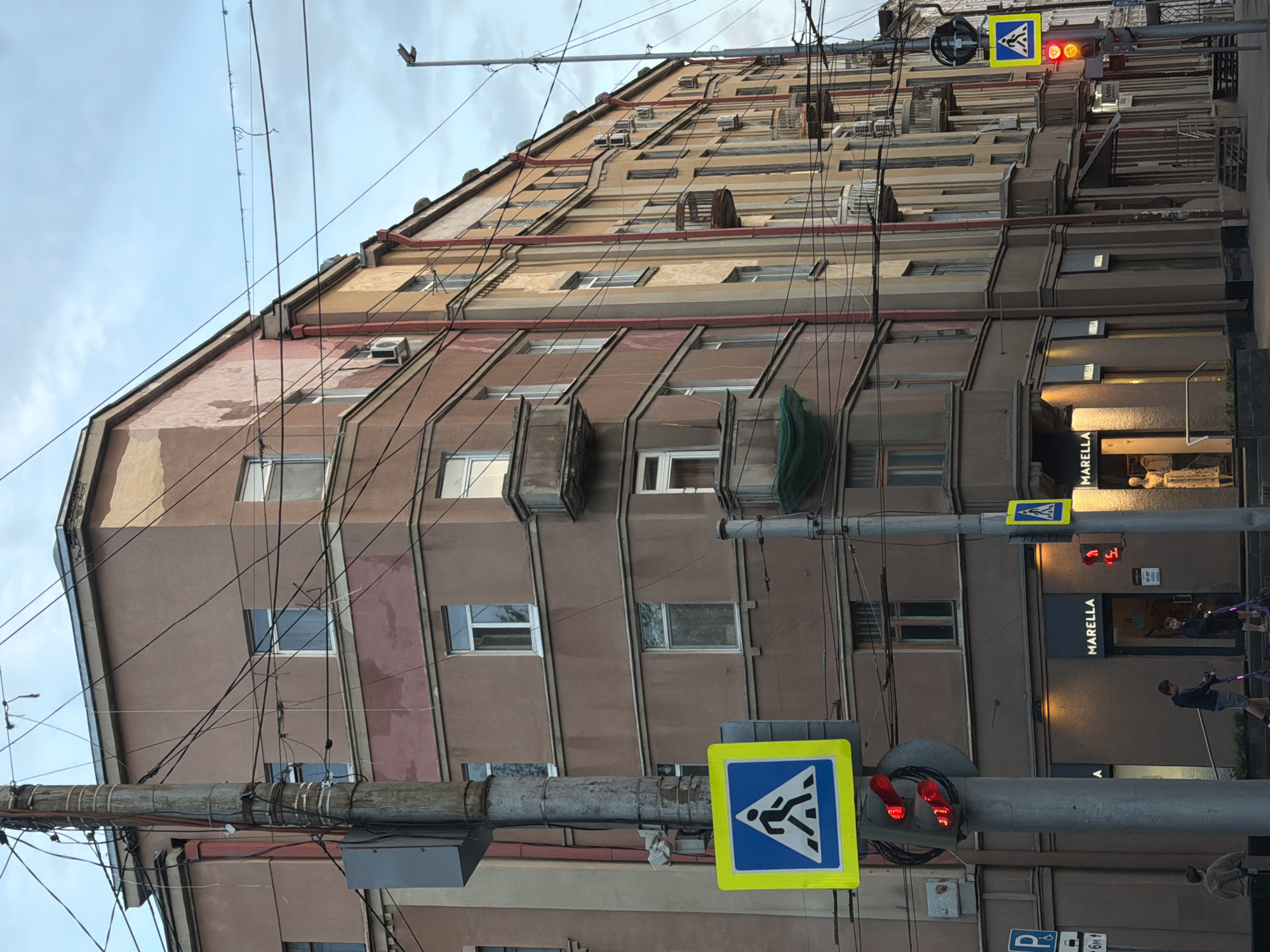 The image depicts a street scene in Saratov, Russia, showcasing a multi-story building and a street intersection. The building has a textured facade with windows, balconies, and external fixtures. Utility poles and a dense network of wires are prominent in the foreground. Two pedestrian crossing signs are visible, mounted on traffic light poles. The traffic lights are red. Street level businesses include a "Marella" store. People are walking on the sidewalk. The sky is overcast.