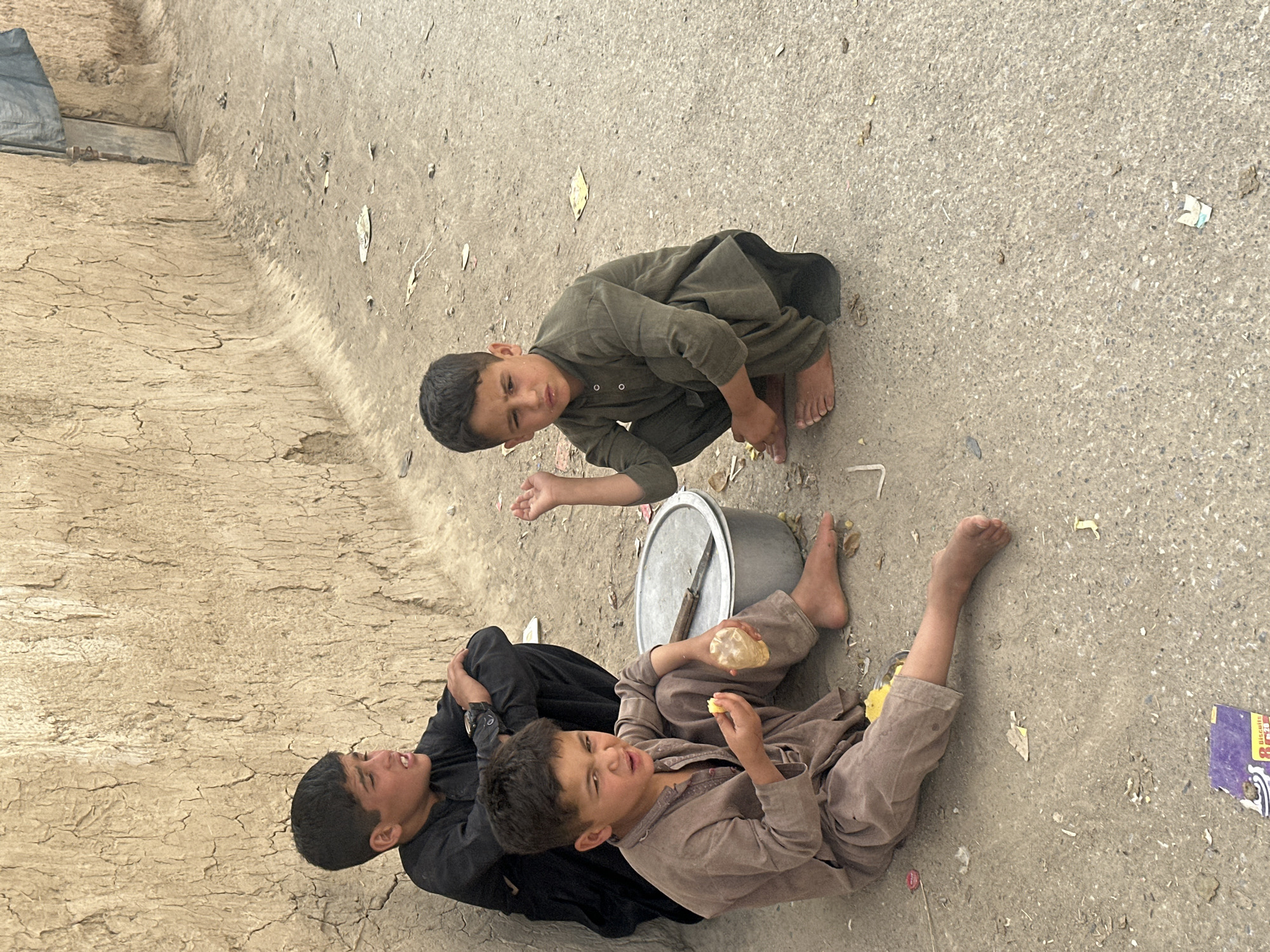 Three barefoot boys sit on a dirt and concrete surface in Quetta City Tehsil, Pakistan. One boy, dressed in dark green, squats with his right hand extended. Another boy, wearing brown clothing, lies on his back with a piece of bread in each hand. A third boy, wearing black clothing, sits beside him. A metal pot with a knife lies between them. Debris is scattered on the ground. A mud wall is on the left.