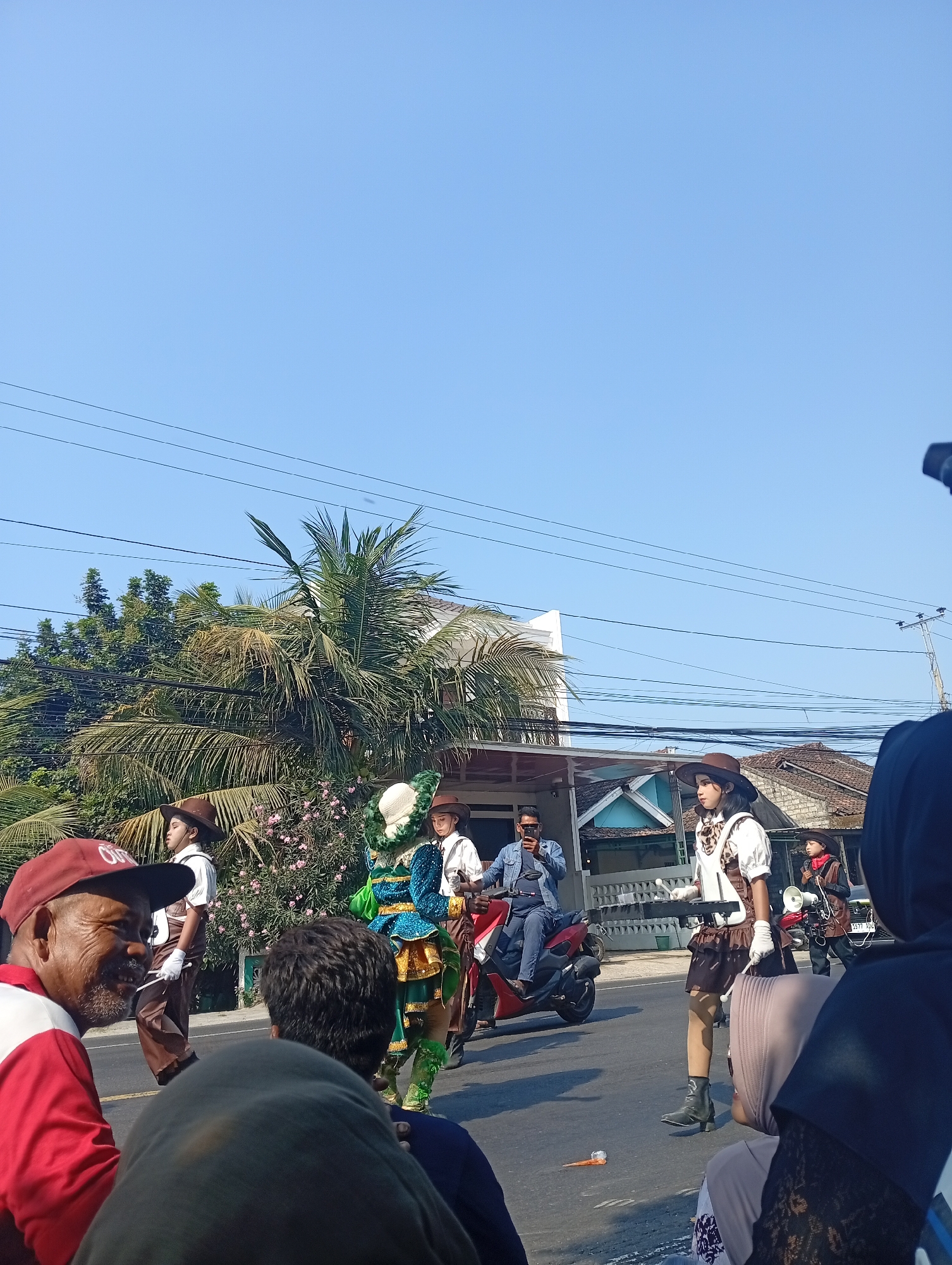 In Sarang, Indonesia, a street parade is underway under a clear blue sky. Several people are walking in costumes. One individual is dressed in a green and blue elaborate costume, with a white hat. They are surrounded by others in western-style outfits, including cowboy hats, vests, and boots. One person rides a red scooter, while another stands next to it, holding a smartphone. Spectators are visible in the foreground, some wearing headscarves. In the background, houses and trees line the street, with power lines overhead. The overall atmosphere appears festive and communal.