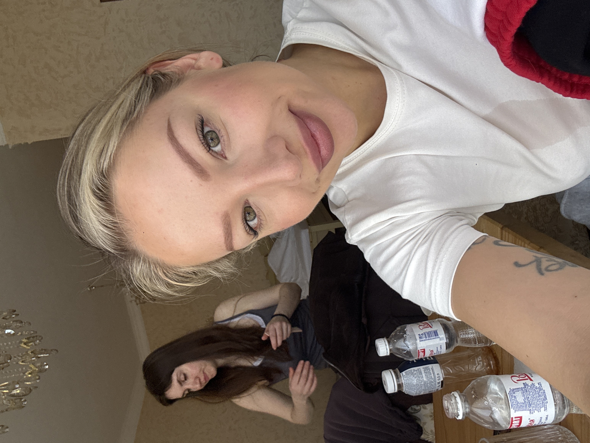 The image depicts a selfie taken by a blonde woman with fair skin, light eyes, and visible lip color. She is wearing a white t-shirt with a red and black garment partially visible. Her left arm is extended, revealing a tattoo. Several empty plastic water bottles are arranged on a wooden surface to her right.
In the background, reflected in a mirrored surface, is another woman with long dark hair, wearing a tank top. She appears to be seated or lying down, with her hands visible. A chandelier is partially visible in the upper left corner of the reflection.
The image does not provide any visual evidence to confirm the location as Baksan, Russia.
