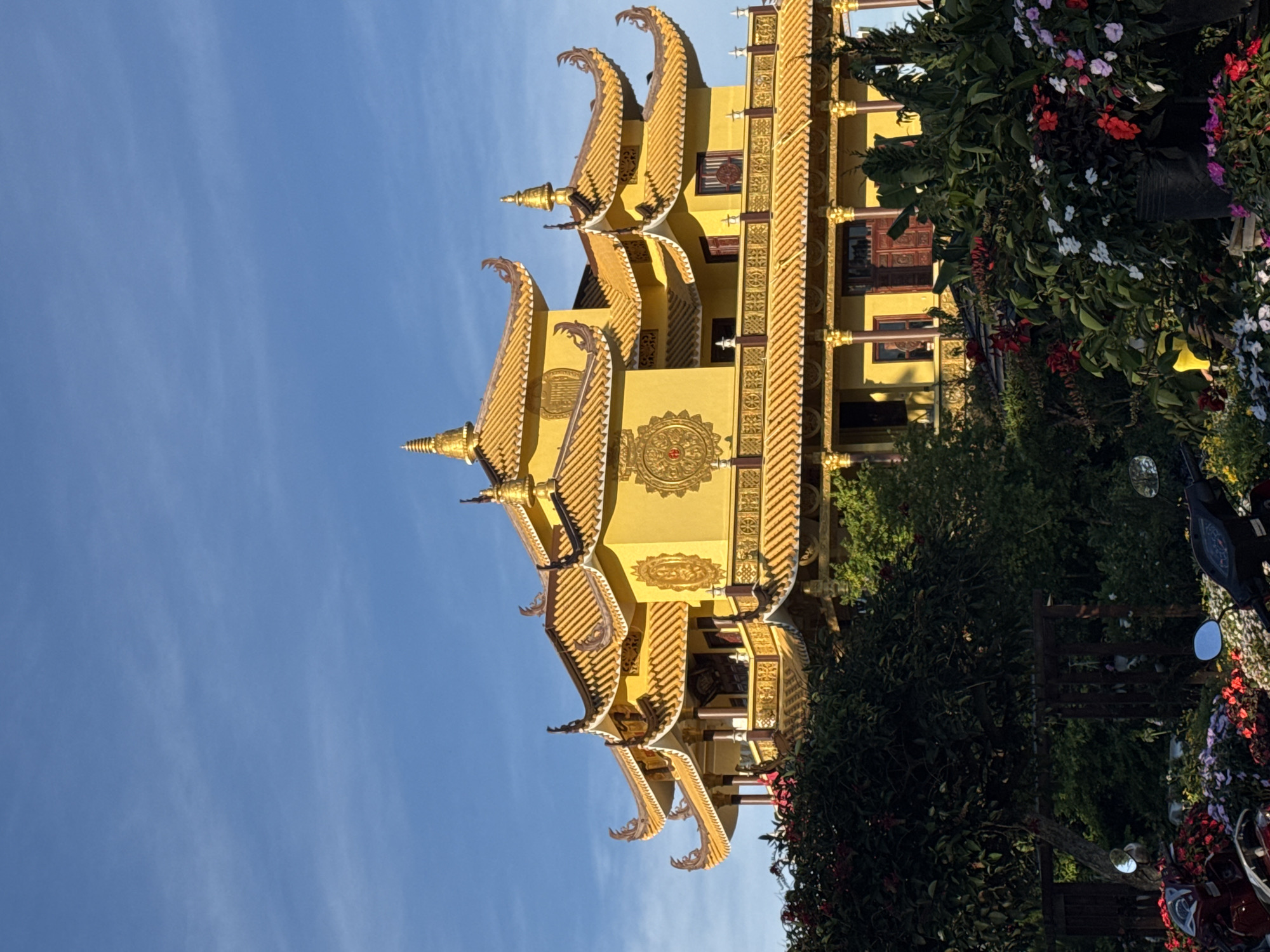 The image depicts the Van Hanh Pagoda in Da Lat, Vietnam. The multi-tiered, yellow and gold structure is prominently featured against a bright blue sky. The architecture includes ornate roof details with pointed finials. Foliage, including flowering plants in red, white, and purple hues, is visible on the right side of the image, partially obscuring the base of the building. A dark-colored motorbike is also visible amidst the foliage. The pagoda is a testament to the Buddhist influence in the region.