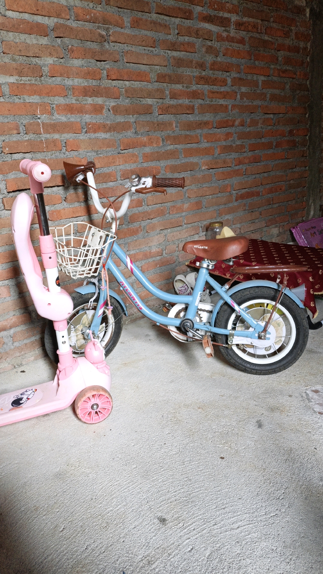 A pink children's scooter and a light blue children's bicycle are parked next to each other on a concrete floor against a red brick wall. The scooter is on the left, with its handlebars and front wheel visible. The bicycle is on the right, with its frame, wheels, seat, and handlebars clearly seen. The bicycle has a basket on the front and a seat on the back. Behind the bicycle is a patterned cloth covering something. The scene appears to be an outdoor or semi-outdoor location, possibly a yard or alleyway. The location is described as Cikal, Indonesia.