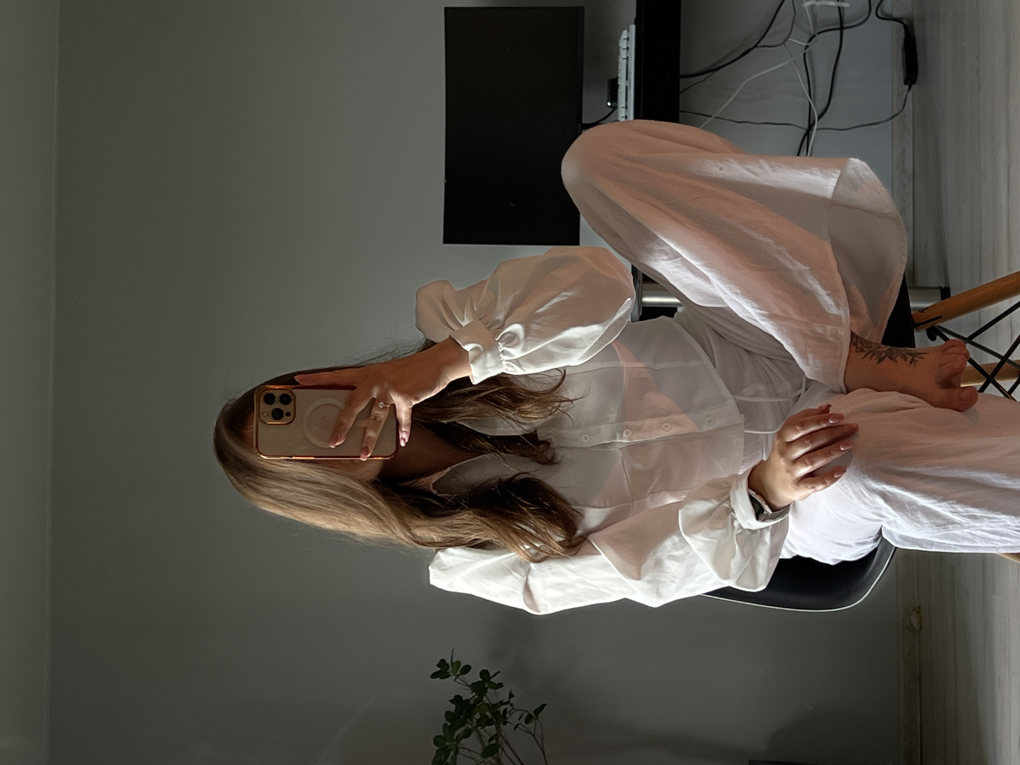 A person is seated indoors, located in Ternopil, Ukraine. The individual has long brown hair and is dressed in a white collared shirt featuring puffed sleeves and white wide-leg pants. A gold-framed smartphone with a circular beige accessory on its case is held by the right hand, obscuring the person's face. The right hand exhibits a light pink fingernail and a ring on the ring finger. A smartwatch is worn on the left wrist, and the left hand rests on the right knee. A tattoo is visible on the left foot. The person occupies a black chair with wooden legs. To the right, a black desk supports an unlit black computer monitor and a white keyboard; multiple black cables and a power adapter are visible beneath the desk. A green potted plant is partially visible on the left side of the frame. The immediate environment consists of a plain gray wall and a light-colored floor.