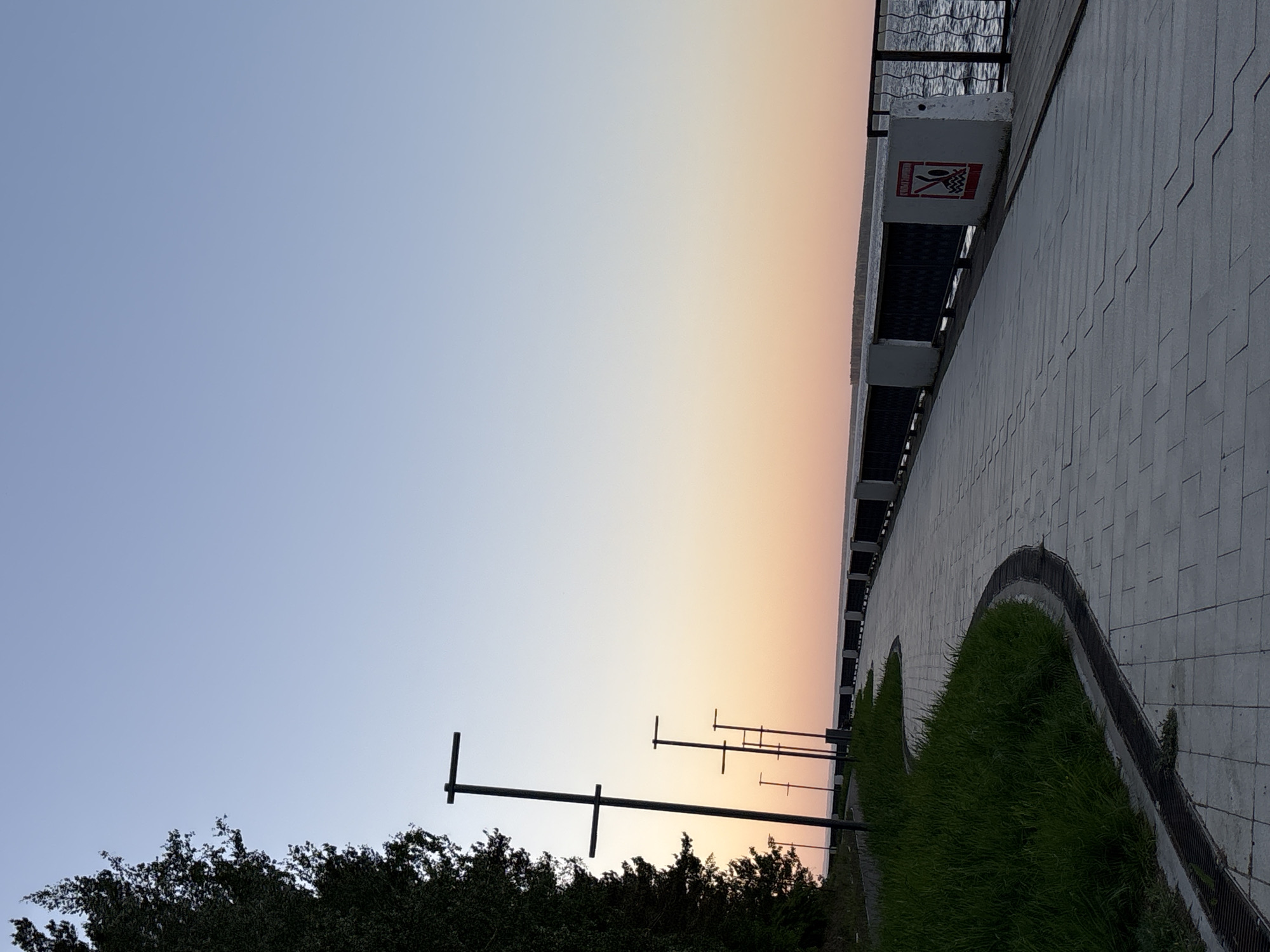 The image shows a sunset view in Chaikovsky, Russia. The sky transitions from blue to orange, illuminating a waterfront promenade. The promenade is paved with grey tiles and features a curved green space. A building with a warning sign and attached benches is visible on the right. Several streetlights line the edge of the promenade, and trees are visible along the bottom of the frame. The scene appears deserted.