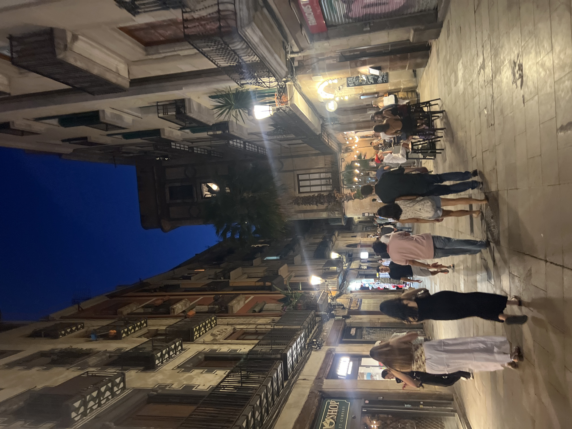 The image shows a street scene in Barcelona, Spain, at twilight. The street is lined with buildings with balconies and shops on the ground floor. People are walking on the cobblestone street and sitting at tables outside a cafe. A palm tree is visible in the background. The sky is a deep blue, indicating it is evening.