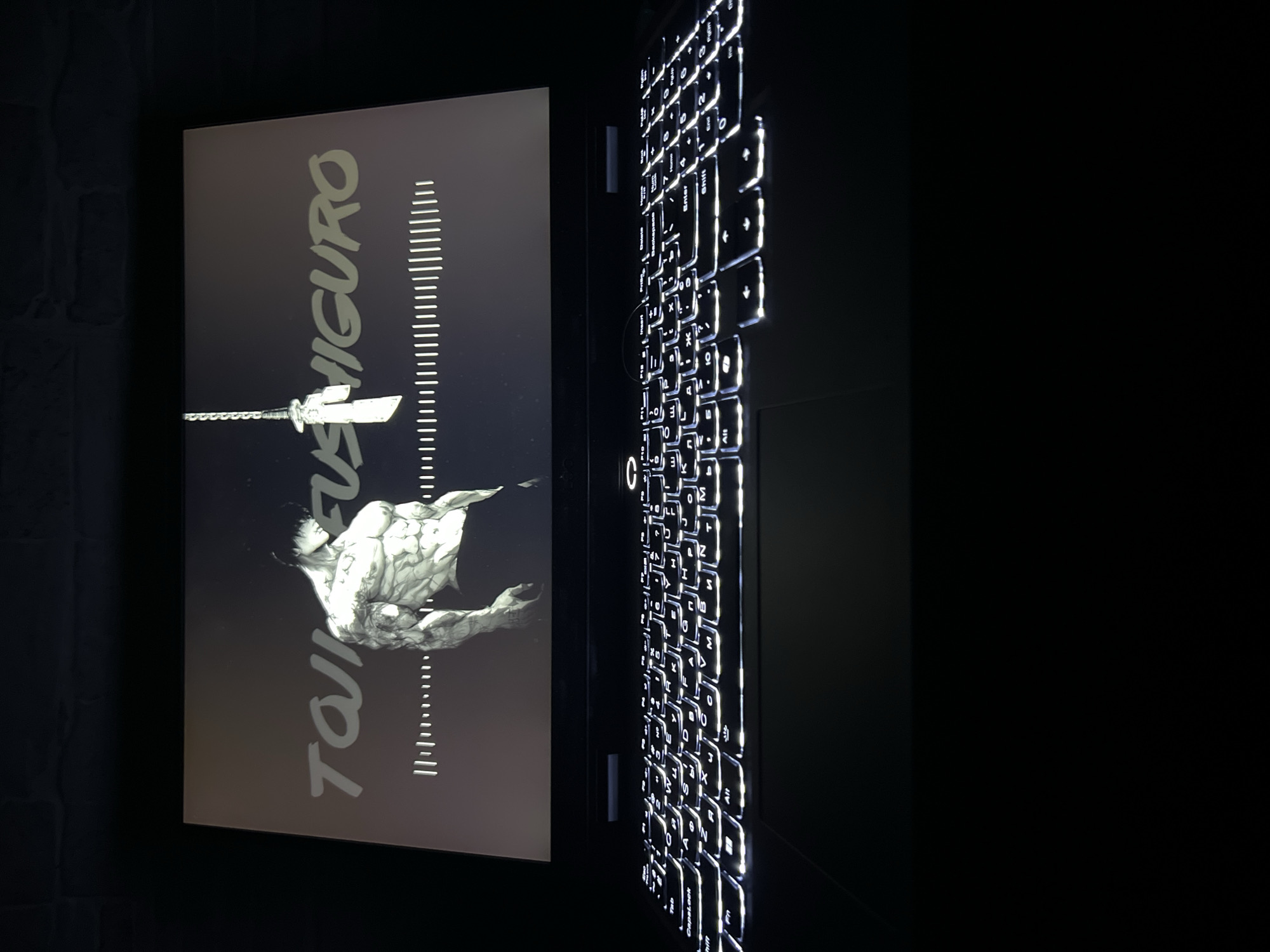 A laptop is positioned on a dark surface, likely a desk or table, with its screen illuminated. The screen displays an image of a muscular, tattooed male figure in a dark, stylized artwork, accompanied by text that appears to read "TOJI FUSHIGURO" in large, light-colored lettering. Below the text and figure, a waveform or equalizer graphic is visible. The laptop's keyboard is also lit up, revealing its keys. The background is dark, with a hint of a textured surface, possibly brick or stone, visible at the top. The overall scene is dimly lit, with the primary light source being the laptop screen and keyboard. There is no indication of a specific location in Sobishchytsi, Ukraine, or any other city.