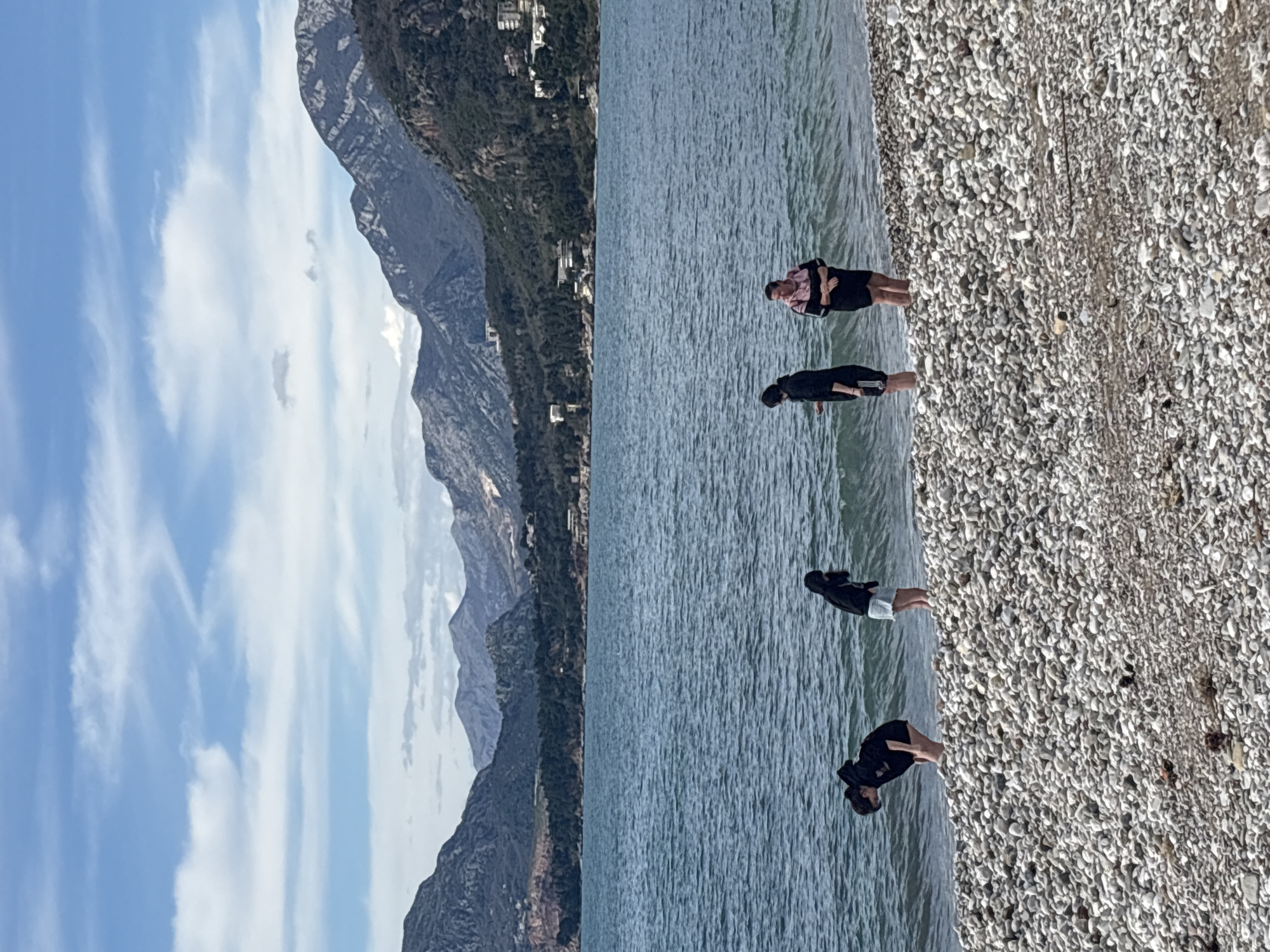 Four people are standing in the shallow water on a pebble beach. Behind them is the Adriatic Sea and a mountain range. Buildings can be seen on the lower slopes of the mountain. The sky is blue with some white clouds. The image was taken in Bar, Montenegro.