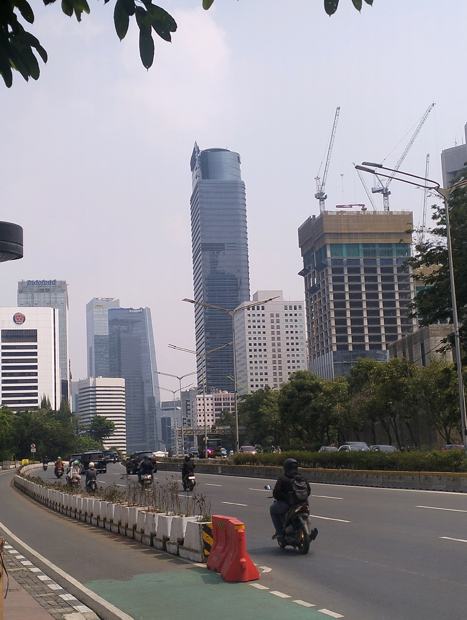 The image depicts a street scene in Central Jakarta, Indonesia. A multi-lane road with a green bike lane curves through the foreground, flanked by planters and concrete barriers. Several motorcycles are in motion on the road, with riders wearing helmets and backpacks. In the background, a cluster of modern high-rise buildings dominates the skyline, including a tall, slender skyscraper with a glass facade and several other office buildings, some of which appear to be under construction with cranes visible. Trees and foliage line the sides of the road, interspersed with streetlights. The sky is overcast, with some green leaves from an overhanging tree visible at the top of the frame.