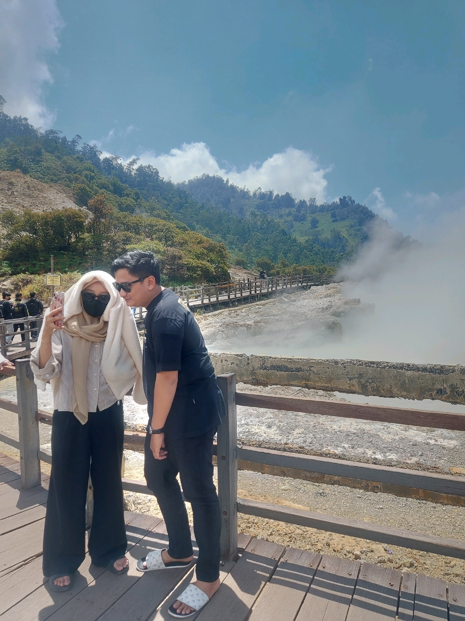 This image captures a couple at the Kawah Sikidang crater in Dieng Plateau, Wonosobo Regency, Central Java, Indonesia. The couple stands on a wooden walkway overlooking the volcanic crater, which is emitting a significant amount of white steam. The man, wearing a black t-shirt and sunglasses, leans in towards the woman. The woman, dressed in a striped shirt, black pants, a hijab, and a face mask, holds up her phone to take a selfie. She is also draped with a light-colored shawl or blanket. The background features lush green hills and a clear blue sky with scattered clouds. The walkway appears to be constructed of wood and is bordered by wooden railings. The crater area itself is rocky and appears to be covered in mineral deposits, with the steam obscuring much of the detail within the crater. Other visitors can be seen on a further section of the wooden walkway in the background. The overall atmosphere suggests a tourist visit to a natural geothermal attraction.