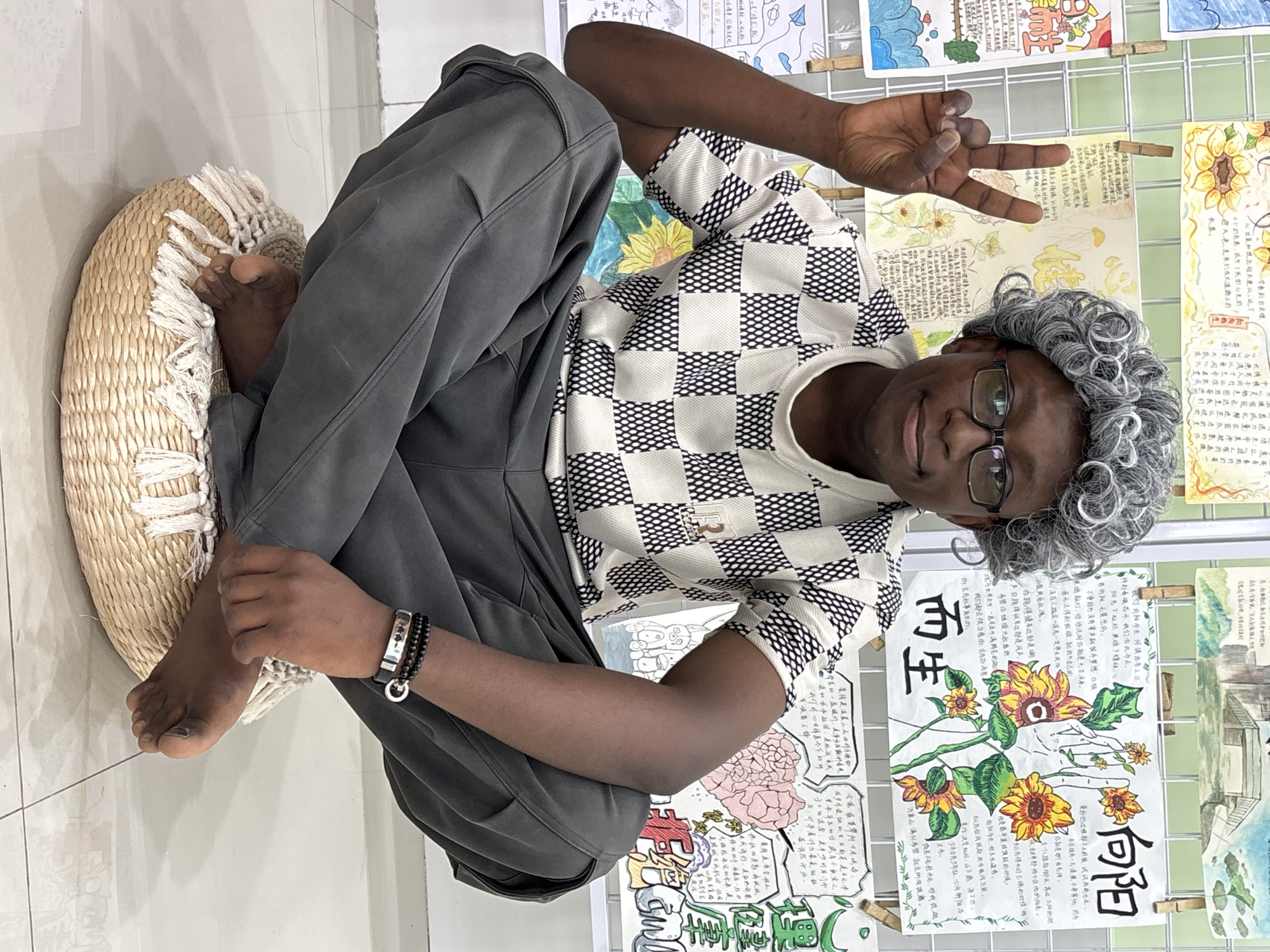A person is sitting on a wicker cushion on a tiled floor, leaning back against a wire grid display of artwork. The person has dark skin, short dark hair, glasses, and is wearing a grey wig. They are wearing a white shirt with a black checkered pattern and grey sweatpants. The person is making a peace sign with their right hand and is holding a book in their left hand. There are two bracelets on their left wrist. The artwork displayed includes colorful paintings of flowers and buildings with Chinese calligraphy. The lighting appears to be natural, and the scene seems to be indoors. The image was taken in the city of Jiangjin, China.