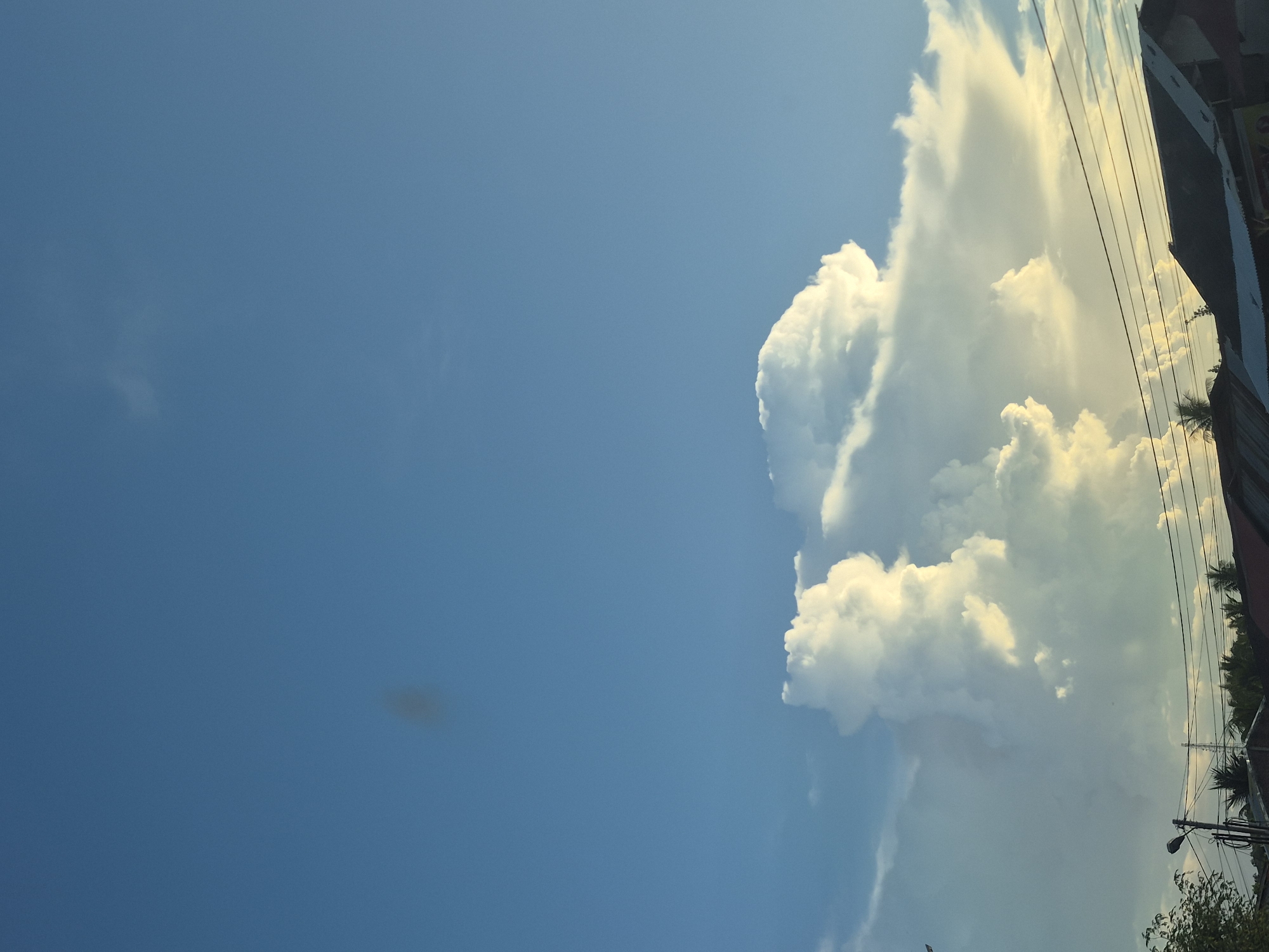 The image captures a view of the sky in Lhokseumawe, Indonesia. A large cumulus cloud formation is present on the right side of the image. The cloud is illuminated by sunlight, giving it a bright, white appearance. To the right, the top portion of a building is visible. Power lines cross in front of the cloud and building. The sky is a clear blue, fading to a brighter shade near the cloud.