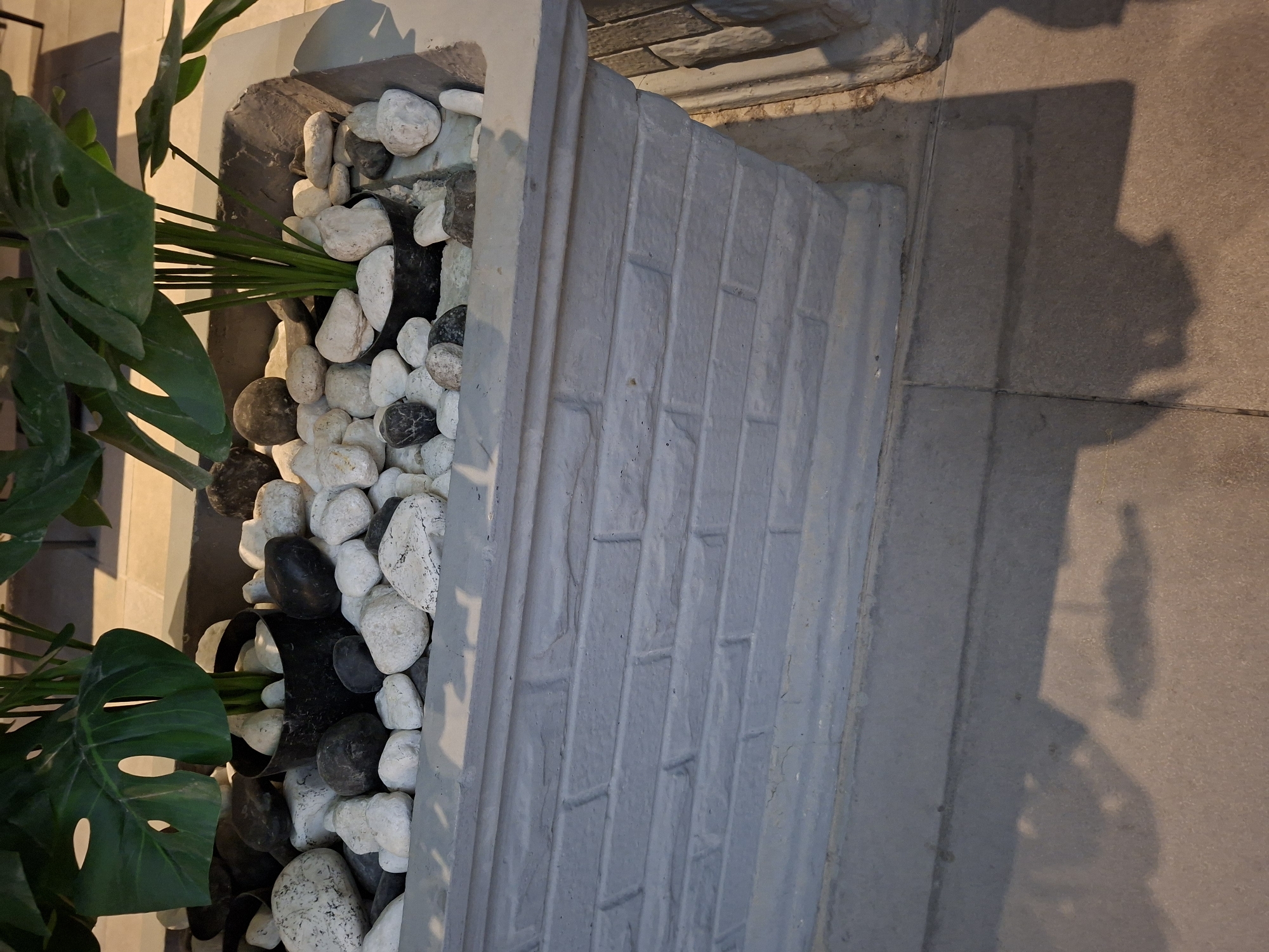 A close-up, eye-level shot focuses on a grey, brick-patterned planter box filled with various sizes of white and dark grey stones. In the center of the planter, a black pot contains a green, artificial monstera plant with large, split leaves and thin stems. The planter is set on a stone-tiled surface where shadows of an unseen object are cast. The background is blurred, showing hints of a wall and possibly outdoor furniture. The scene appears to be in Al Rayyan, Qatar.