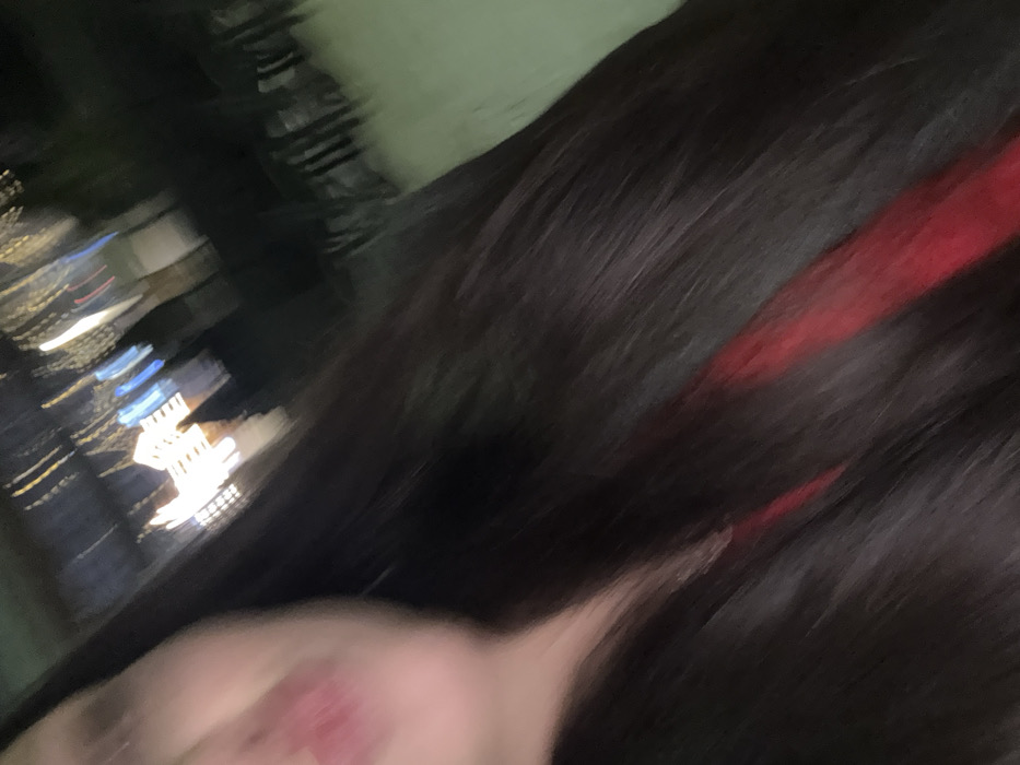 A blurry, angled shot captures a dark-haired person with a streak of red highlights. The background reveals blurred city lights at night, with indistinct buildings. The subject's face is partially visible, showing skin. The overall image has a shaky, out-of-focus quality, suggesting movement or quick capture. The angle and blur make the exact details unclear.