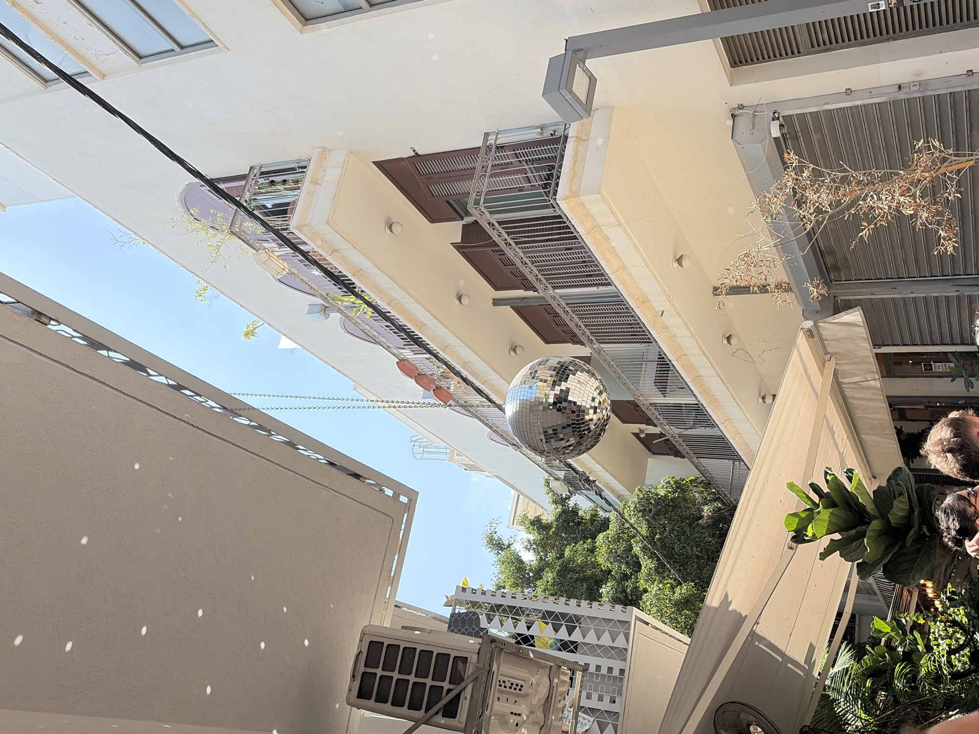 The image shows a disco ball hanging from a wire against a clear blue sky and white building in Tel Aviv, Israel. The building has balconies with railings and dark-colored shutters. Green trees and foliage are visible in the background, as well as parts of an outdoor seating area with a retractable awning and an air conditioning unit. The scene appears to be taken from a street or courtyard, with the camera looking upwards.