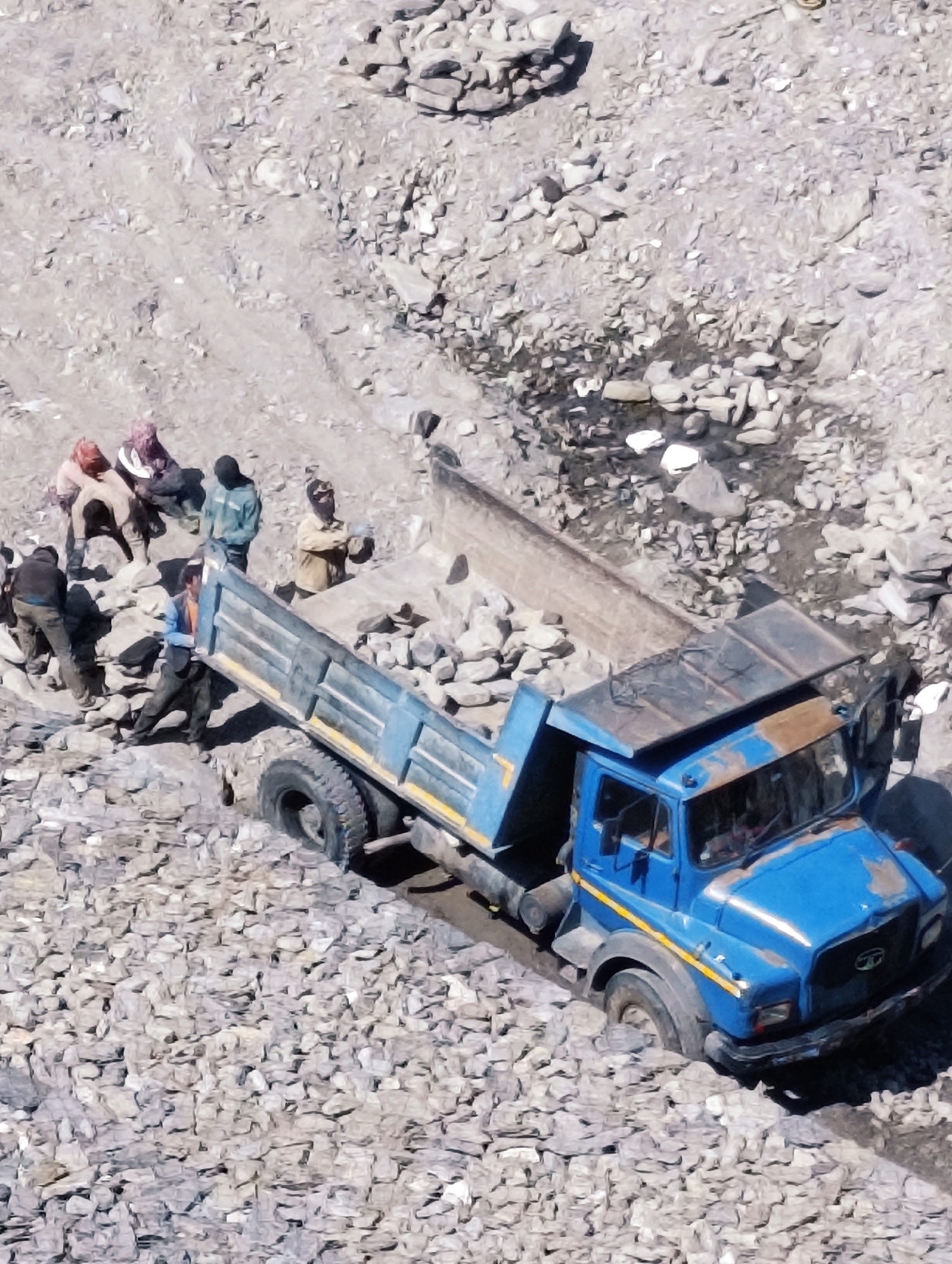 This is an aerial shot of a construction site in Sissu, India, where a blue dump truck is partially submerged in muddy terrain filled with rocks. Several individuals, dressed in casual work attire, are present around the truck, some appearing to be engaged in the loading or unloading process. The environment is rugged and mountainous, characterized by rough ground, loose stones, and a small stream or pool of water. The truck's bed is tilted, indicating an active dumping or loading operation. The overall scene depicts a challenging work environment common in mountainous regions.