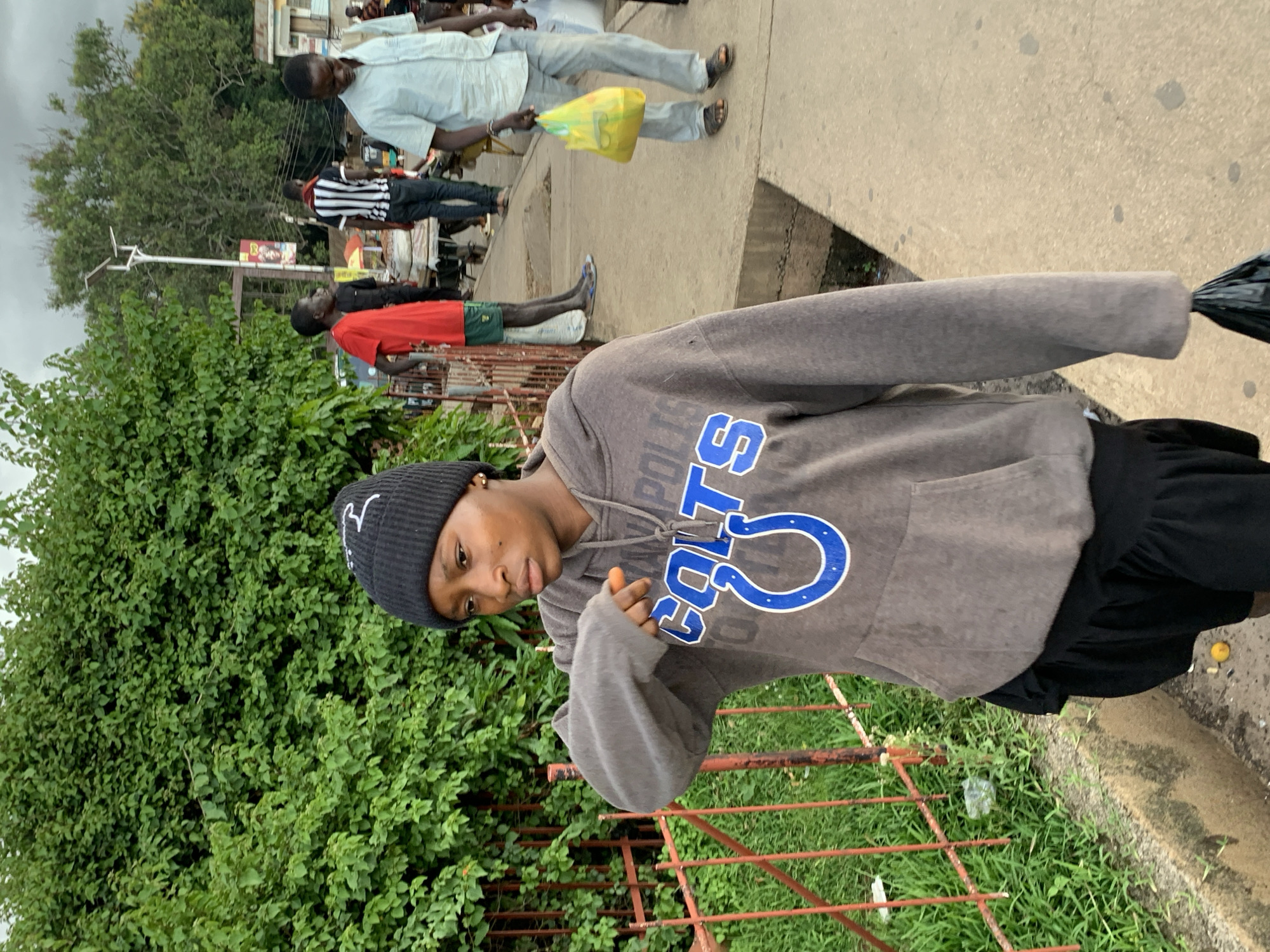 A young Black woman wearing a black beanie and a grey hoodie with "COLTS" written on it, and black shorts walks on a paved sidewalk in Jos, Nigeria. She is carrying a black plastic bag in her right hand. The sidewalk is lined with a red metal fence and a lush green hedge on the left. In the background, other people are walking and standing, including a man in a blue shirt and light-colored pants carrying a yellow plastic bag. A street light with solar panels is visible in the distance, along with a poster on a pole. The sky is overcast.
