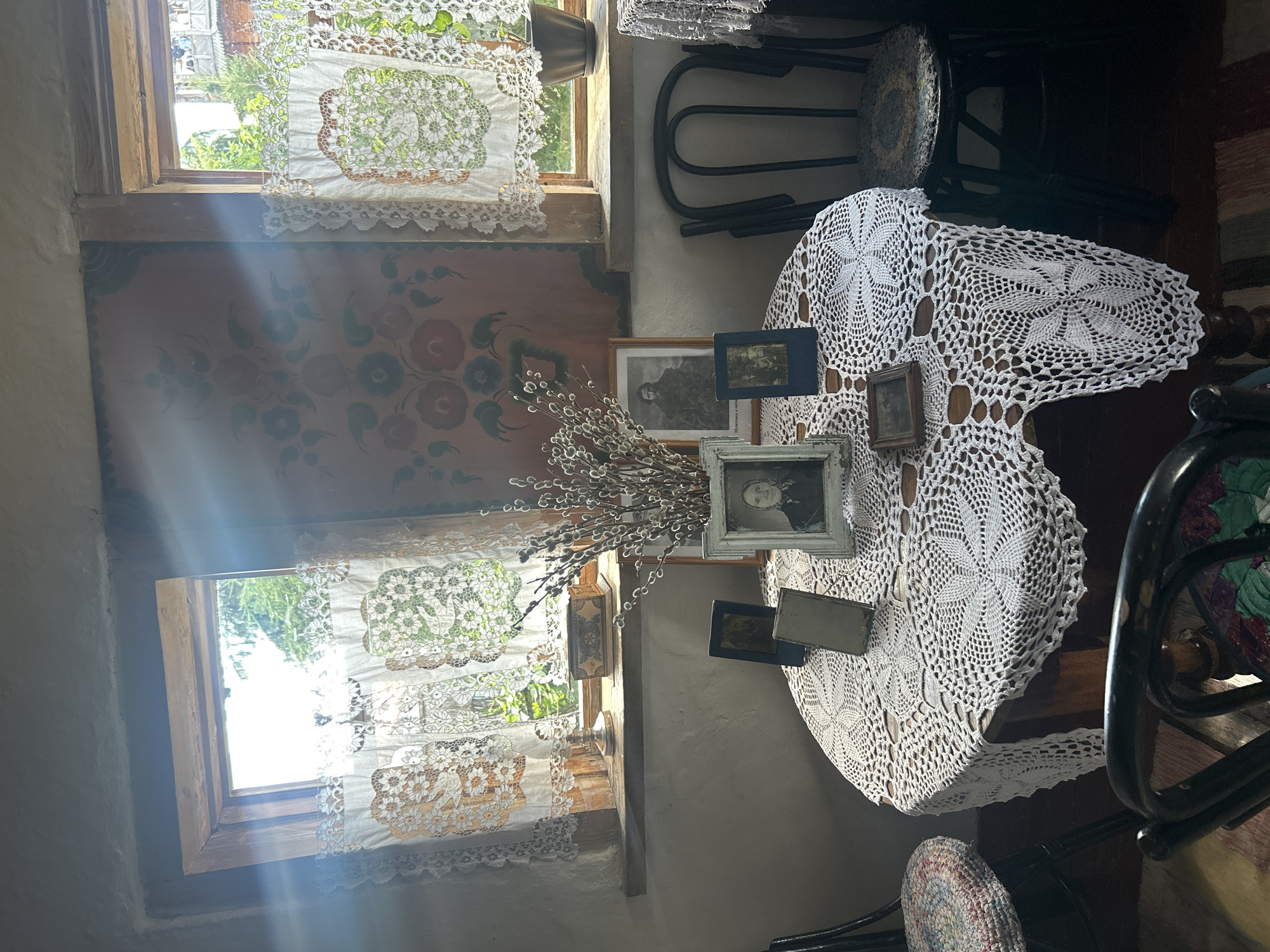 The image depicts an indoor scene, possibly a room in a house, with two windows providing natural light. The room is decorated with what appears to be traditional folk art on the wall between the windows and lace curtains on both windows. A round table is set in the center of the room, covered with a white crocheted tablecloth. On the table are several framed photographs, a vase with dried branches, and other small decorative items. Two chairs are visible, one on the right and a partial view of another on the left. The overall impression is one of a preserved historical or cultural space, perhaps a museum exhibit or a preserved home. The provided context of Bolsherechye, Russia, suggests this could be a scene from a historical dwelling or museum in that location.