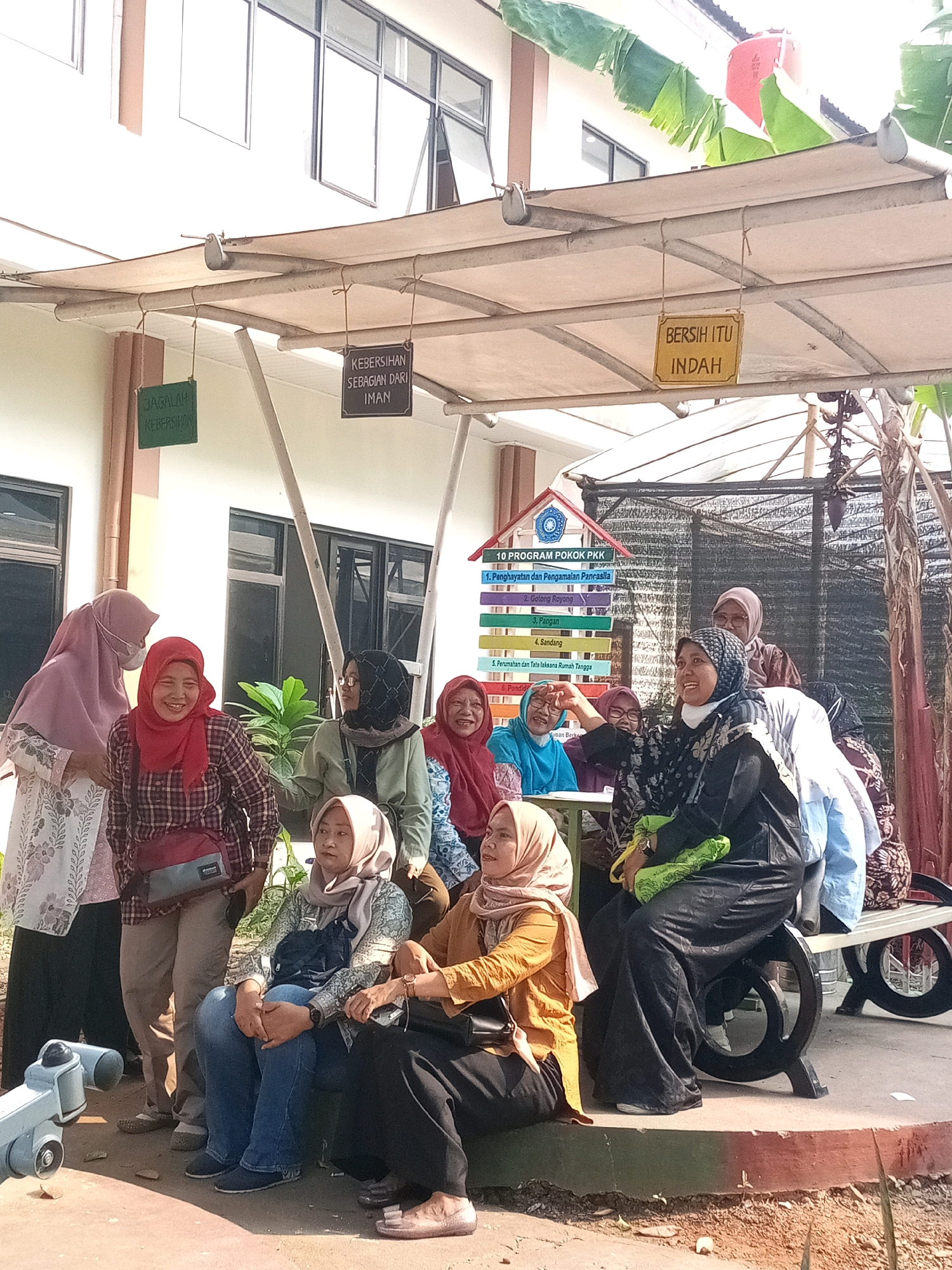 The image shows a group of women posing for a photo outdoors, possibly at a community center or public gathering space in Bekasi, Indonesia. The background features a building with large windows and a covered outdoor area with a beige canopy supported by metal poles. Several signs are visible, including one with "KEBERSIHAN SEBAGIAN DARI IMAN" (Cleanliness is part of faith) and another that says "BERSIH ITU INDAH" (Cleanliness is beautiful). There's also a colorful chart displaying "10 PROGRAM POKOK PKK," which are the 10 core programs of the Family Welfare Movement (PKK) in Indonesia.
The women are dressed in various traditional and modern attire, mostly hijabs and modest clothing. Some are standing, while others are seated on a bench and a low wall. The woman in the foreground, wearing a yellow headscarf and orange jacket, is smiling. Another woman in a red headscarf and plaid shirt is also smiling broadly. The overall atmosphere appears cheerful and social. In the lower left corner, a grey object resembling a metal sculpture or possibly a piece of equipment is visible. The ground is paved with concrete and dirt, with some sparse vegetation. Banana trees are visible in the background on the right side.